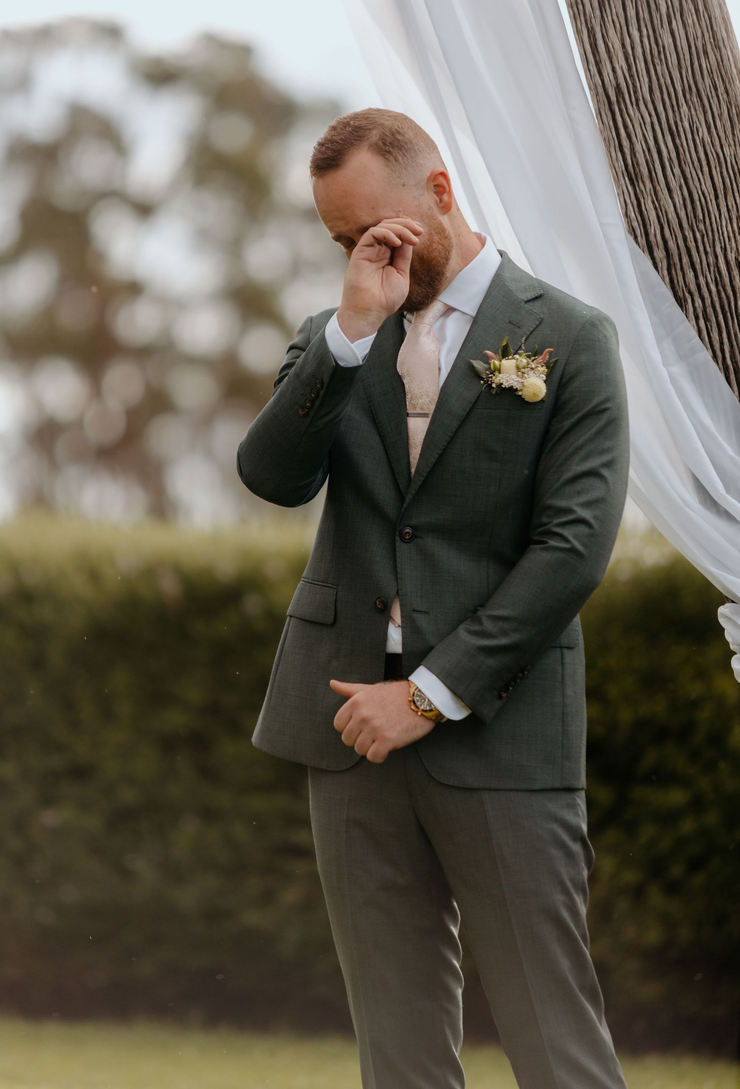 Emotional groom in dark green suit wiping tears at outdoor wedding ceremony with floral boutonniere.