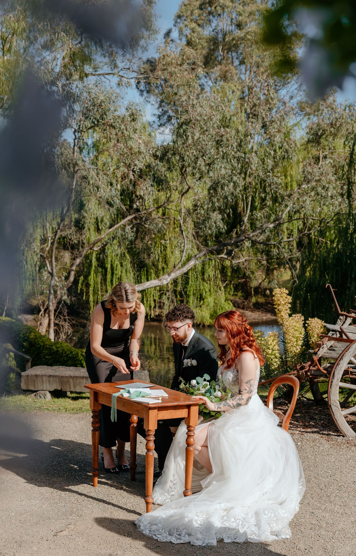Two people in formal wedding attire share a playful moment on wooden benches in a rustic outdoor garden setting.
