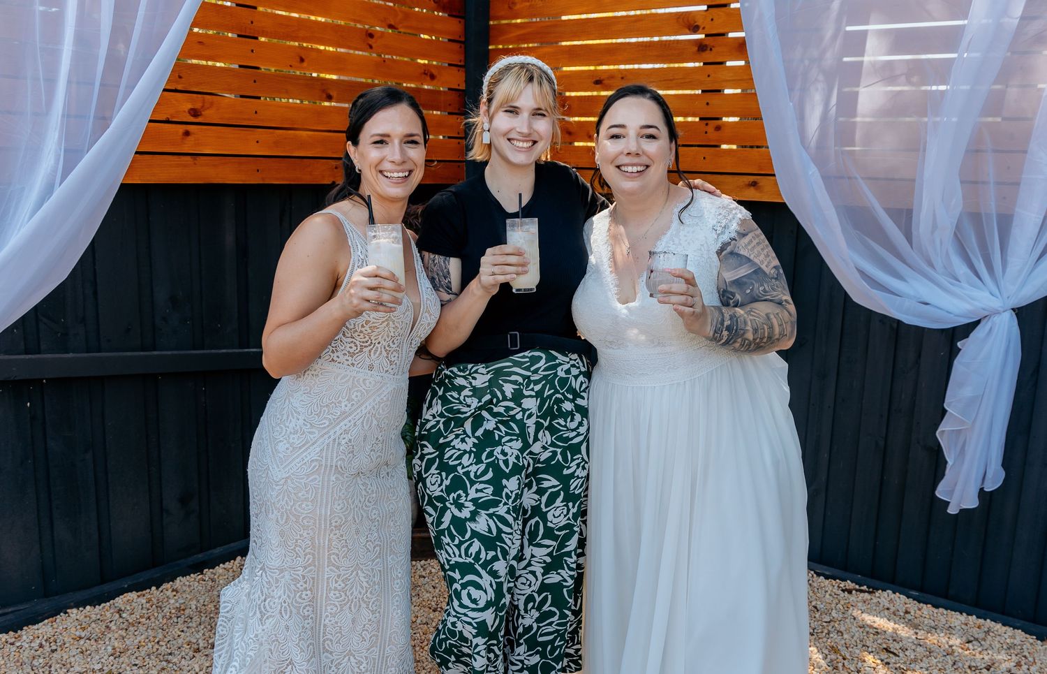 Three people in formal attire smile while holding drinks in front of a wooden wall with white curtains.