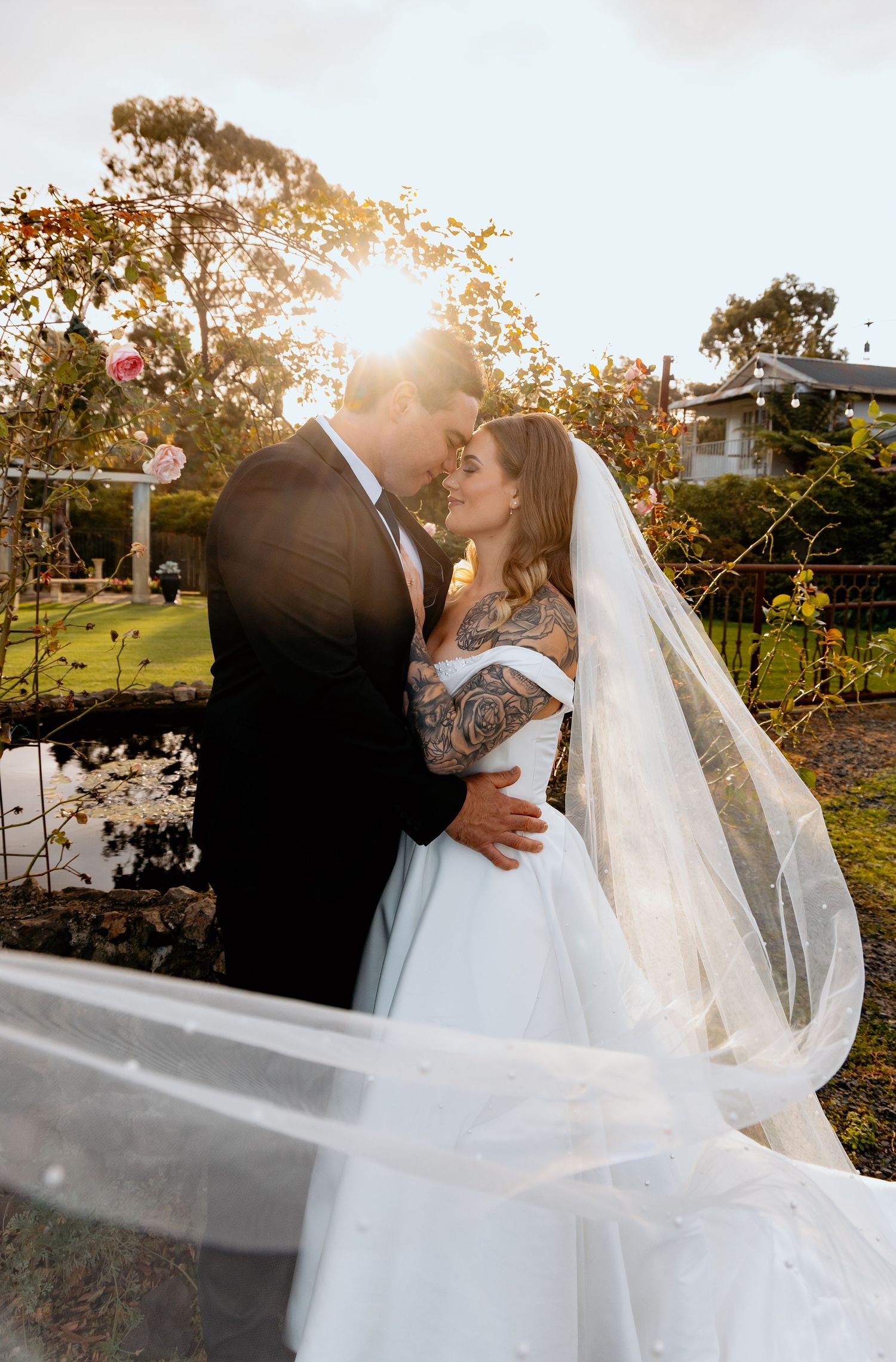 Bride and groom share romantic sunset moment, her flowing veil billowing, in a lush garden setting.