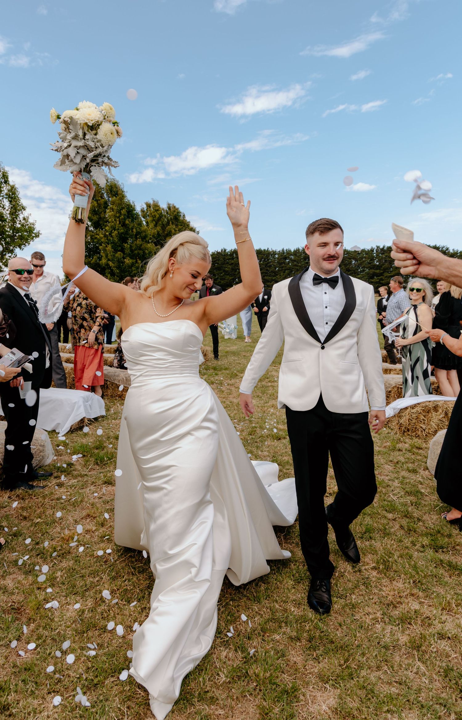 Joyful bride in white strapless gown and groom in white tuxedo celebrate their wedding exit with confetti outdoors.
