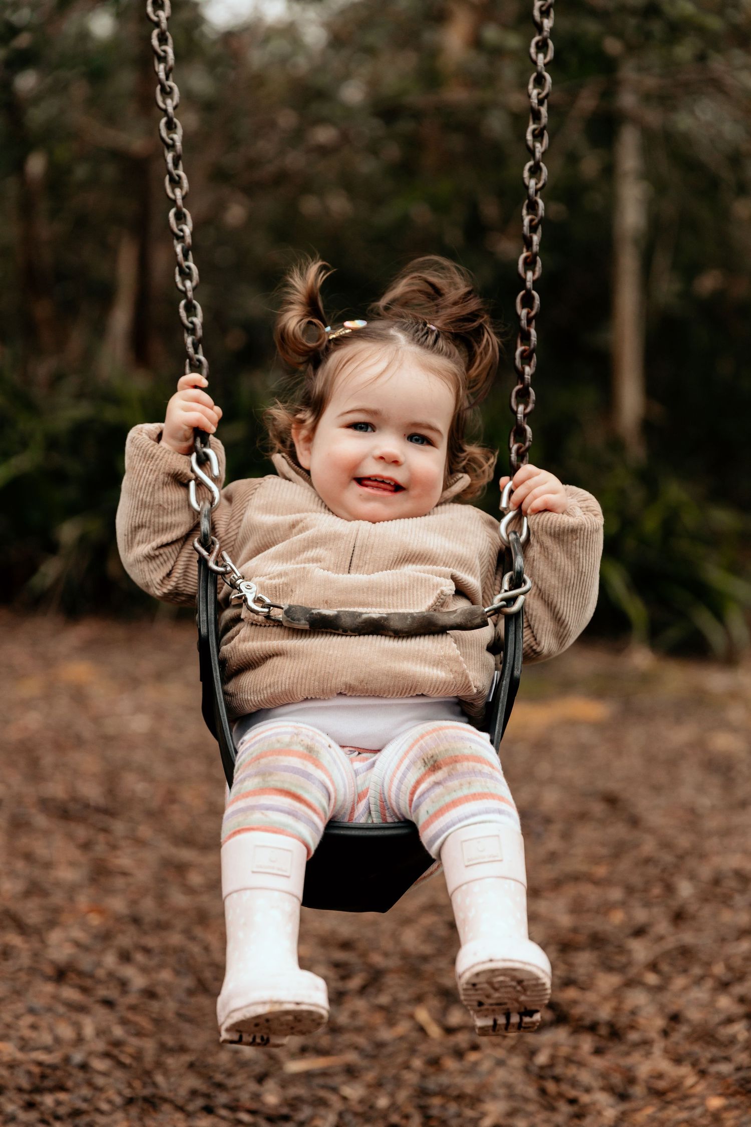 A joyful toddler in a beige sweater swings on a playground swing with a big smile.