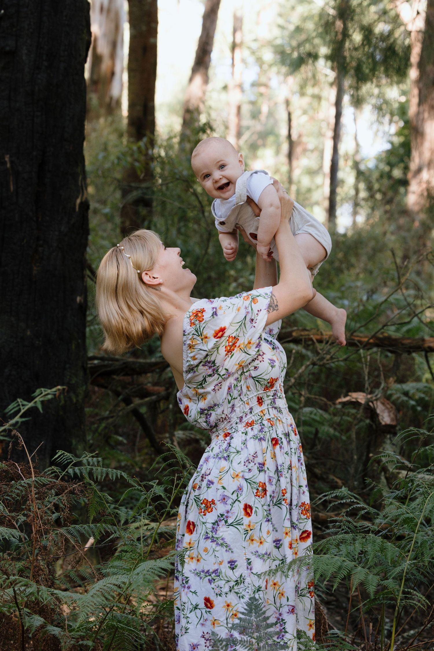 Mother and baby share joyful moment in forest setting wearing floral dress.