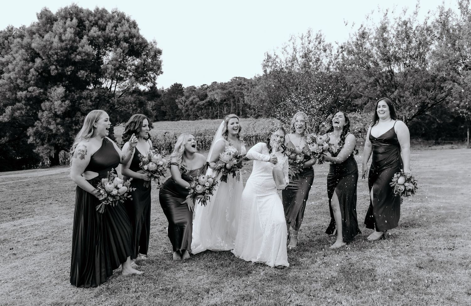 A black and white photo of a bridal party posing together outdoors with bouquets in a natural setting.