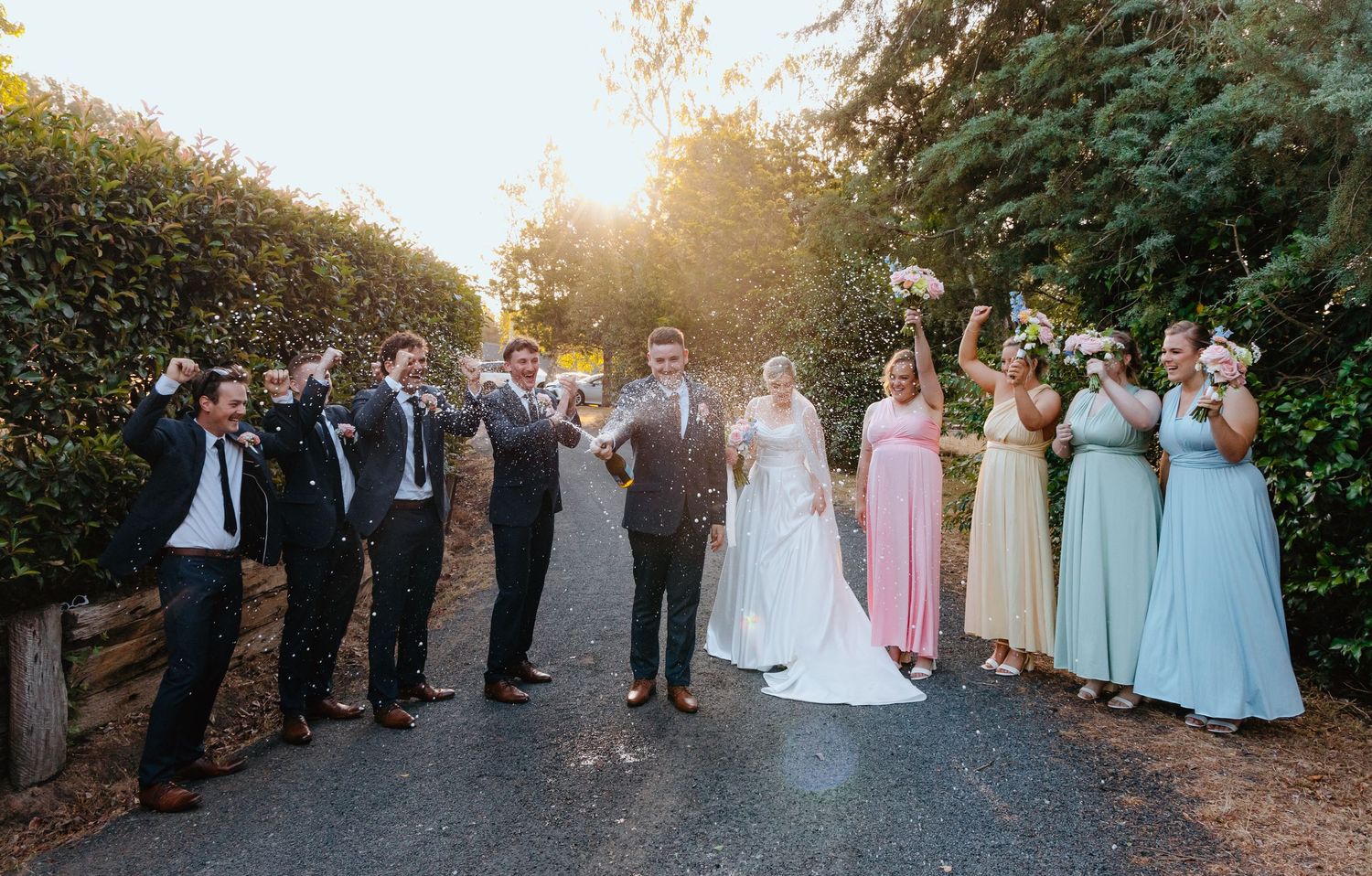 Wedding party celebrating outdoors, bride and groom flanked by groomsmen in suits and bridesmaids in pastel dresses.