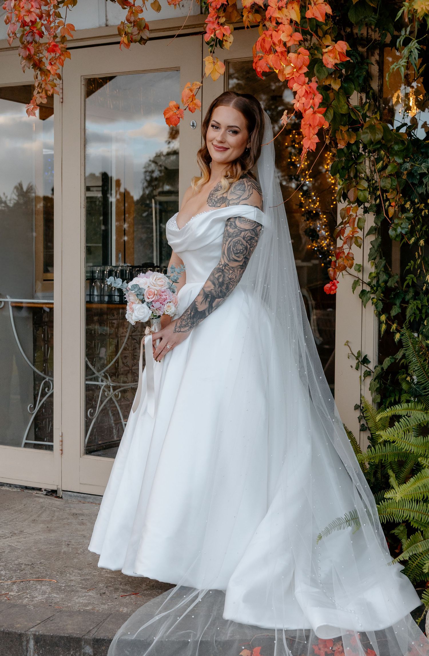 Tattooed bride in off-shoulder white ball gown with veil holds bouquet, posing before autumn foliage backdrop.