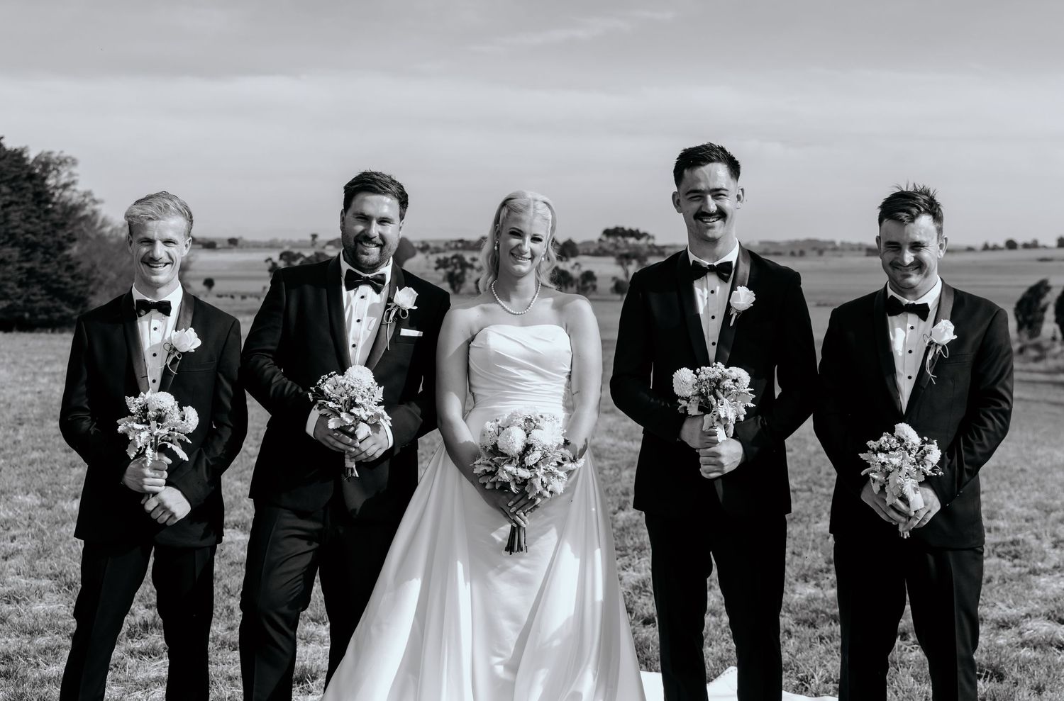 Bride in white gown poses with four groomsmen in black tuxedos holding bouquets in a field.