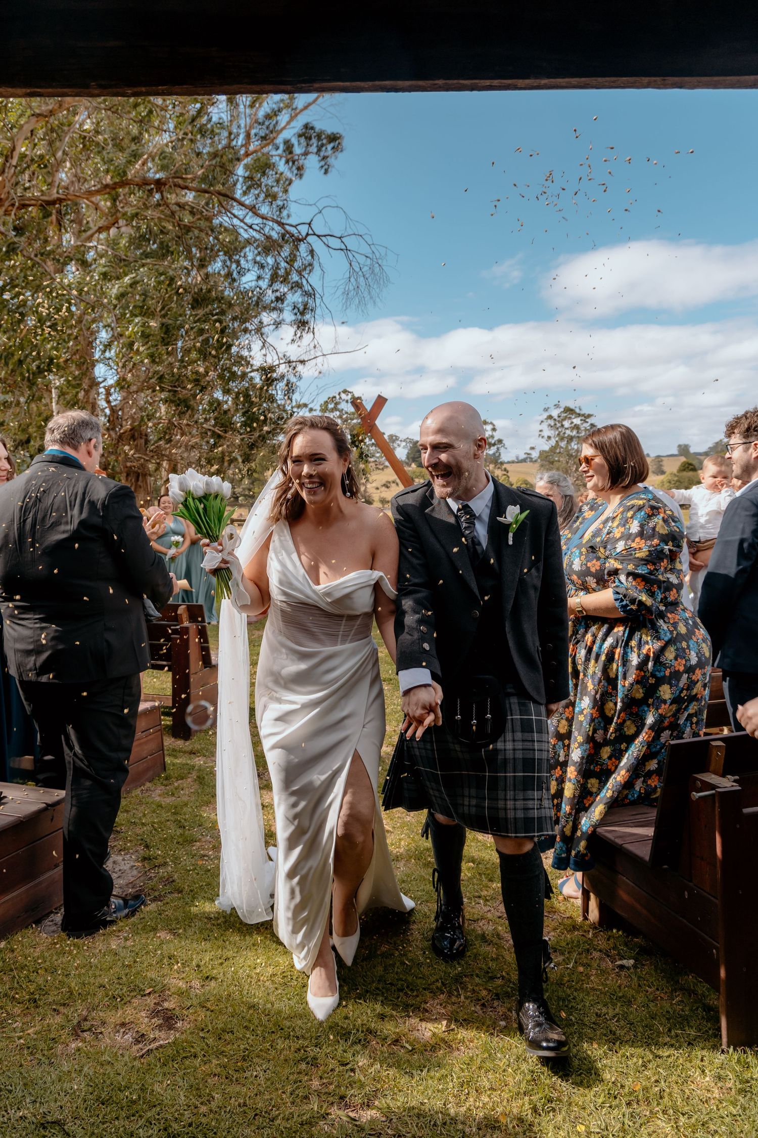 Bride and groom laughing as confetti falls during outdoor wedding ceremony, groom wearing traditional Scottish kilt.