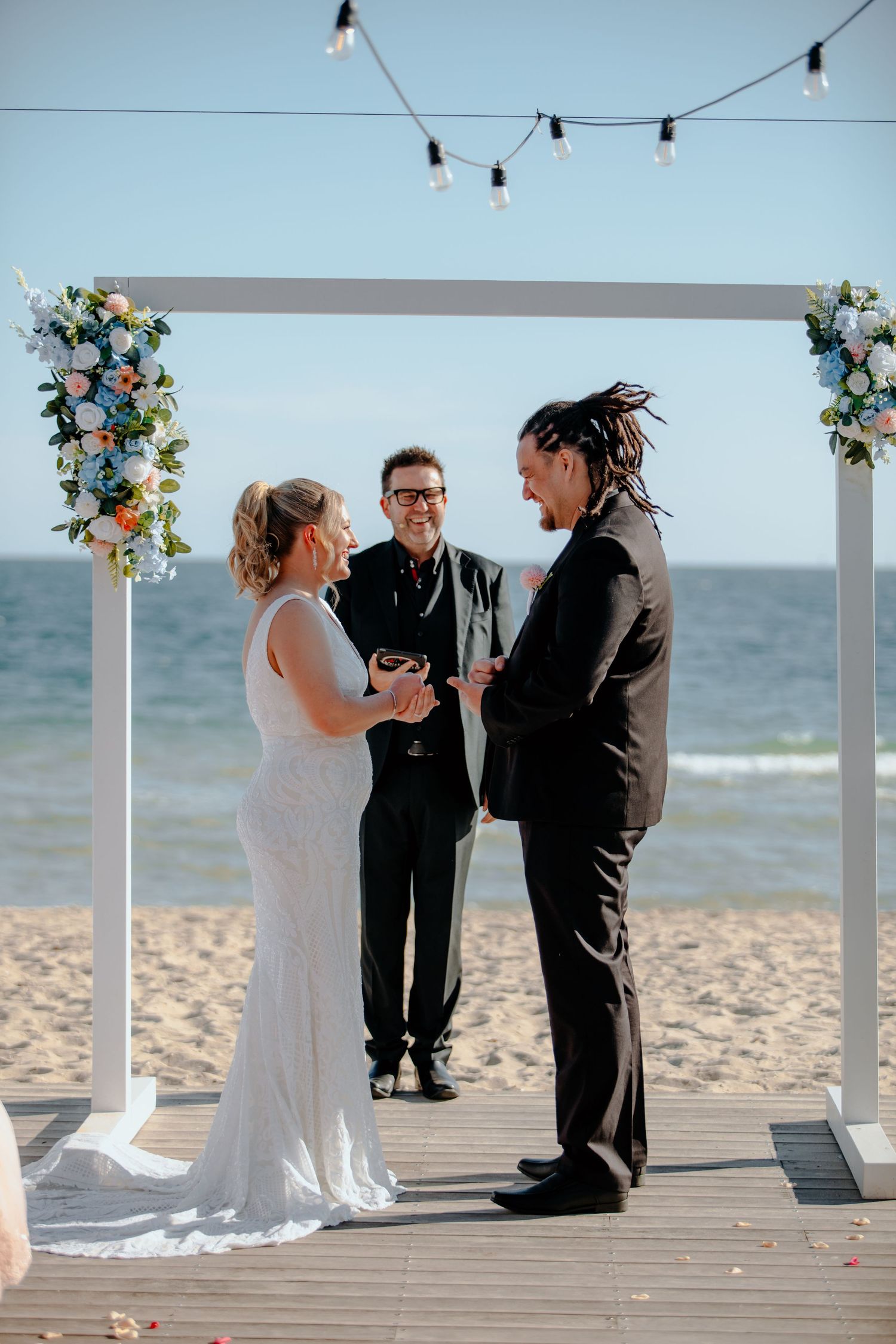 Bride and groom exchange vows under a floral arch on a beachside deck with ocean views.