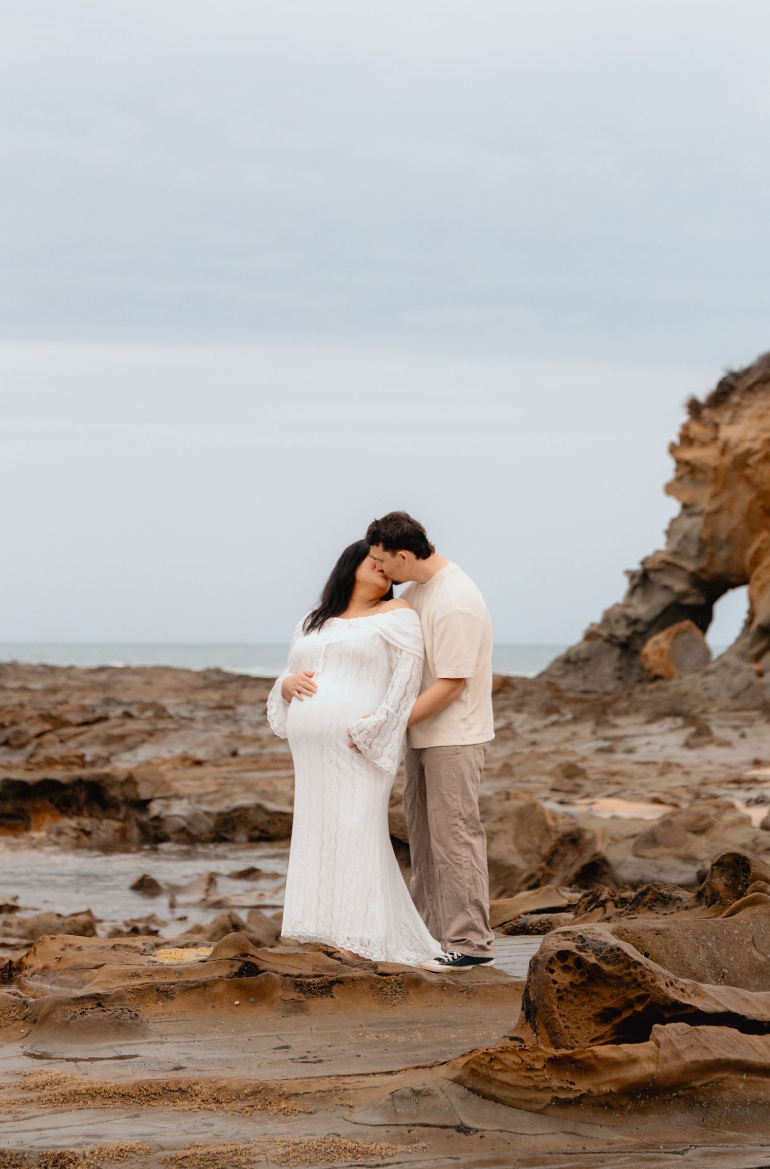 A couple poses together on rocky coastal terrain during sunset for an intimate beach photoshoot.
