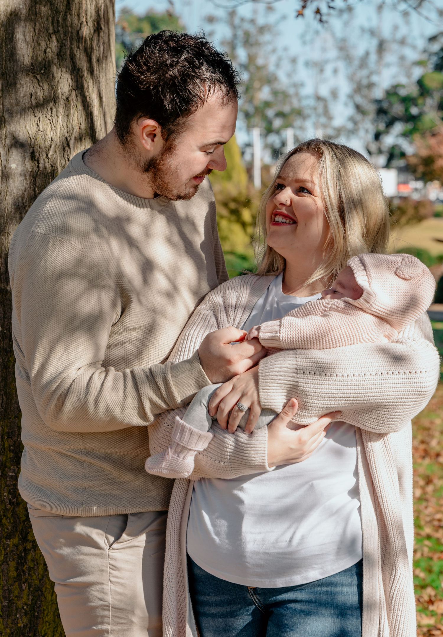 A couple in light-colored sweaters shares a tender moment while holding a baby wrapped in a knit blanket outdoors.