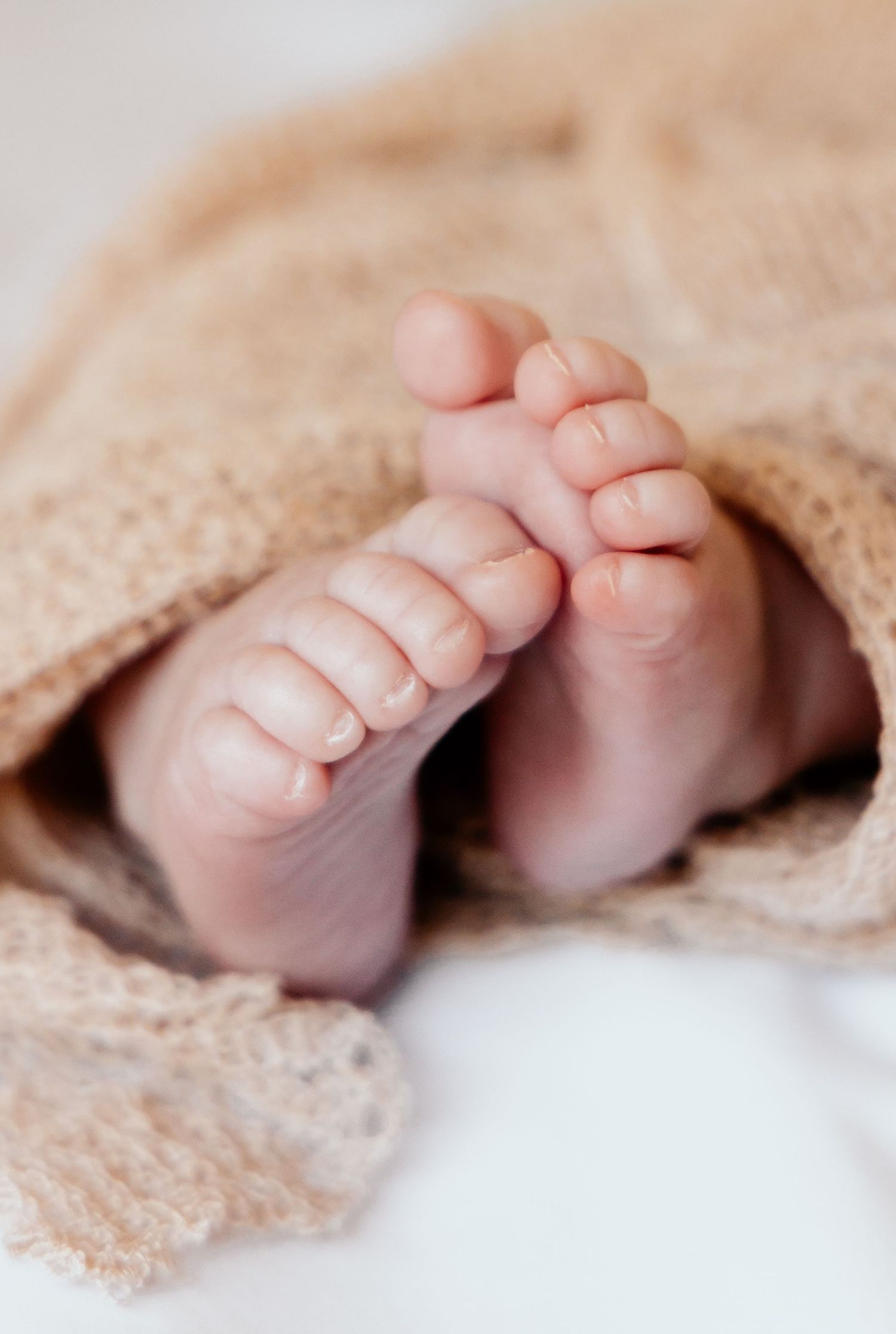 Tiny baby toes peek out from under a soft beige knitted blanket in a tender newborn photograph.