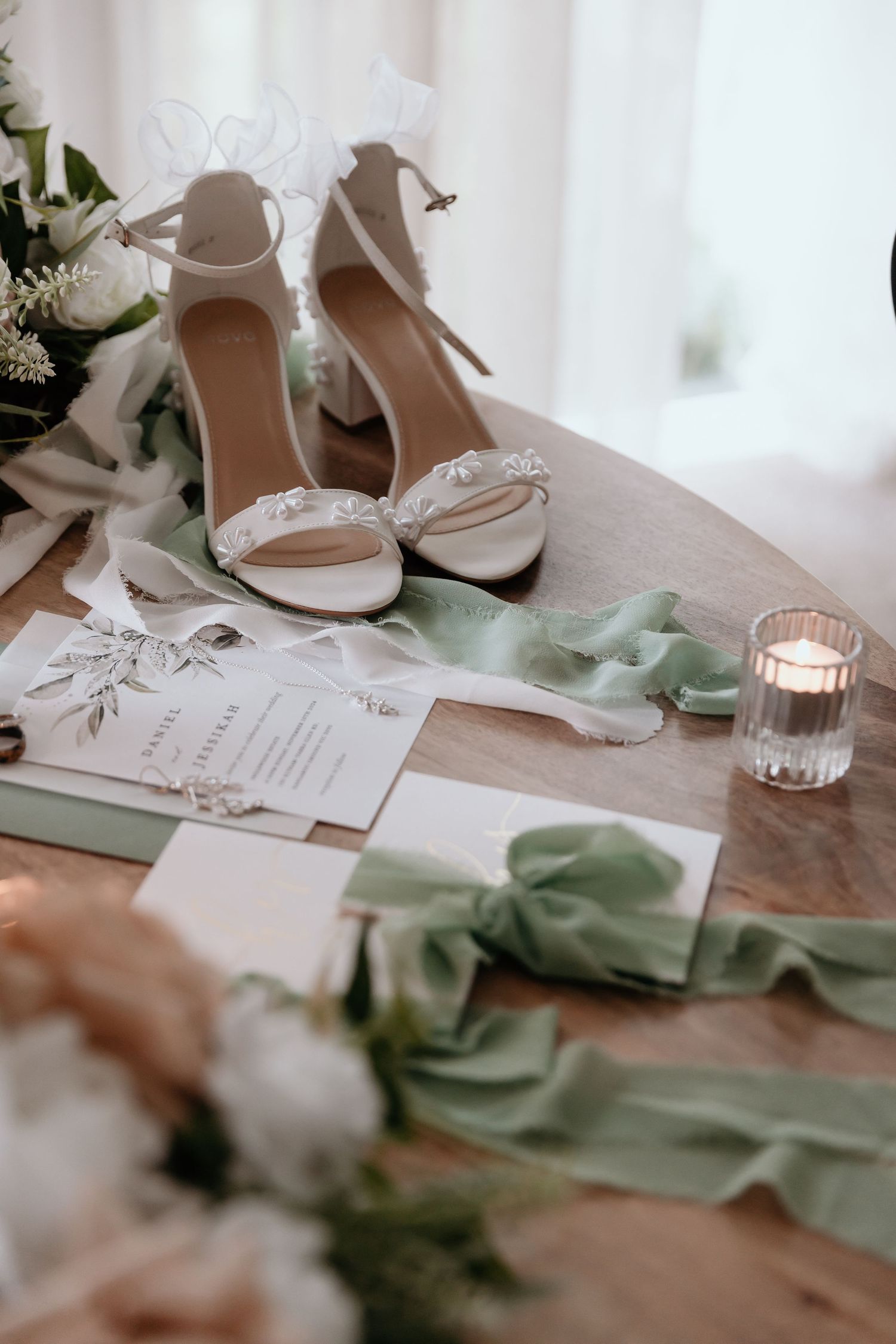 Elegant white wedding shoes and paper stationery arranged with sage green fabric accents and glowing candlelight.