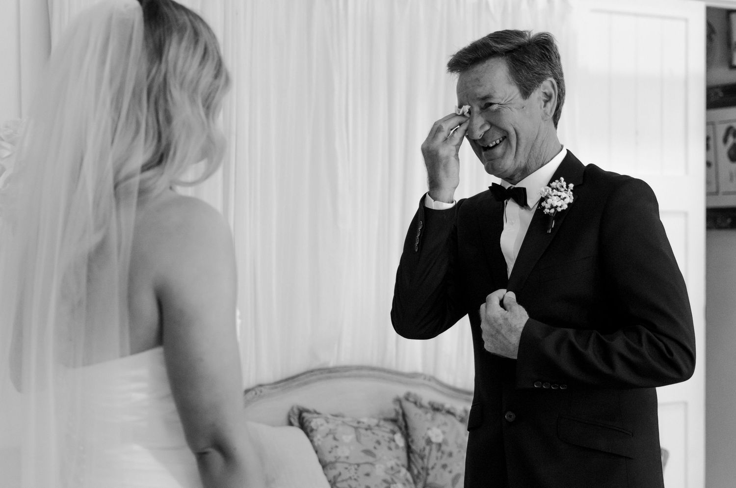 Emotional father wipes tears seeing bride daughter in wedding dress, black and white first look photo.