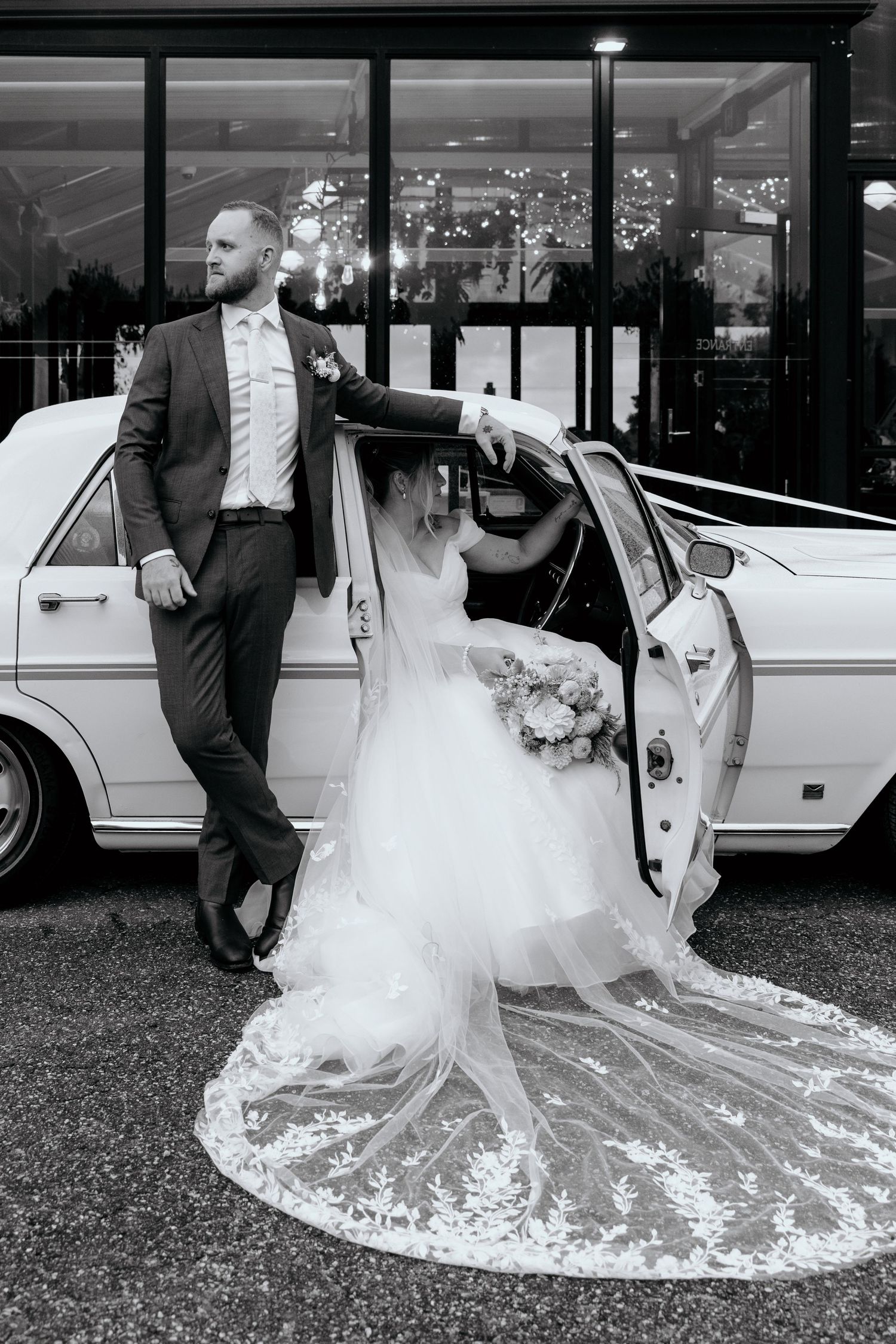 Black and white photo of bride exiting vintage white car as groom stands beside it outside wedding venue.