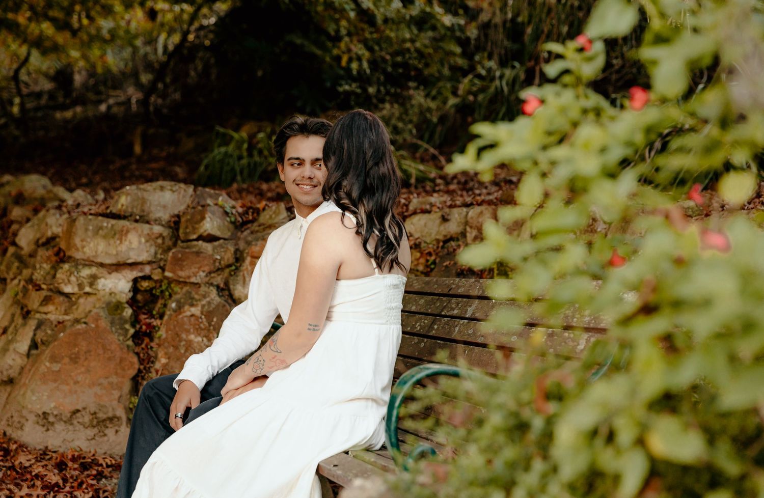 Couple in formal attire sitting on stone wall surrounded by garden foliage.