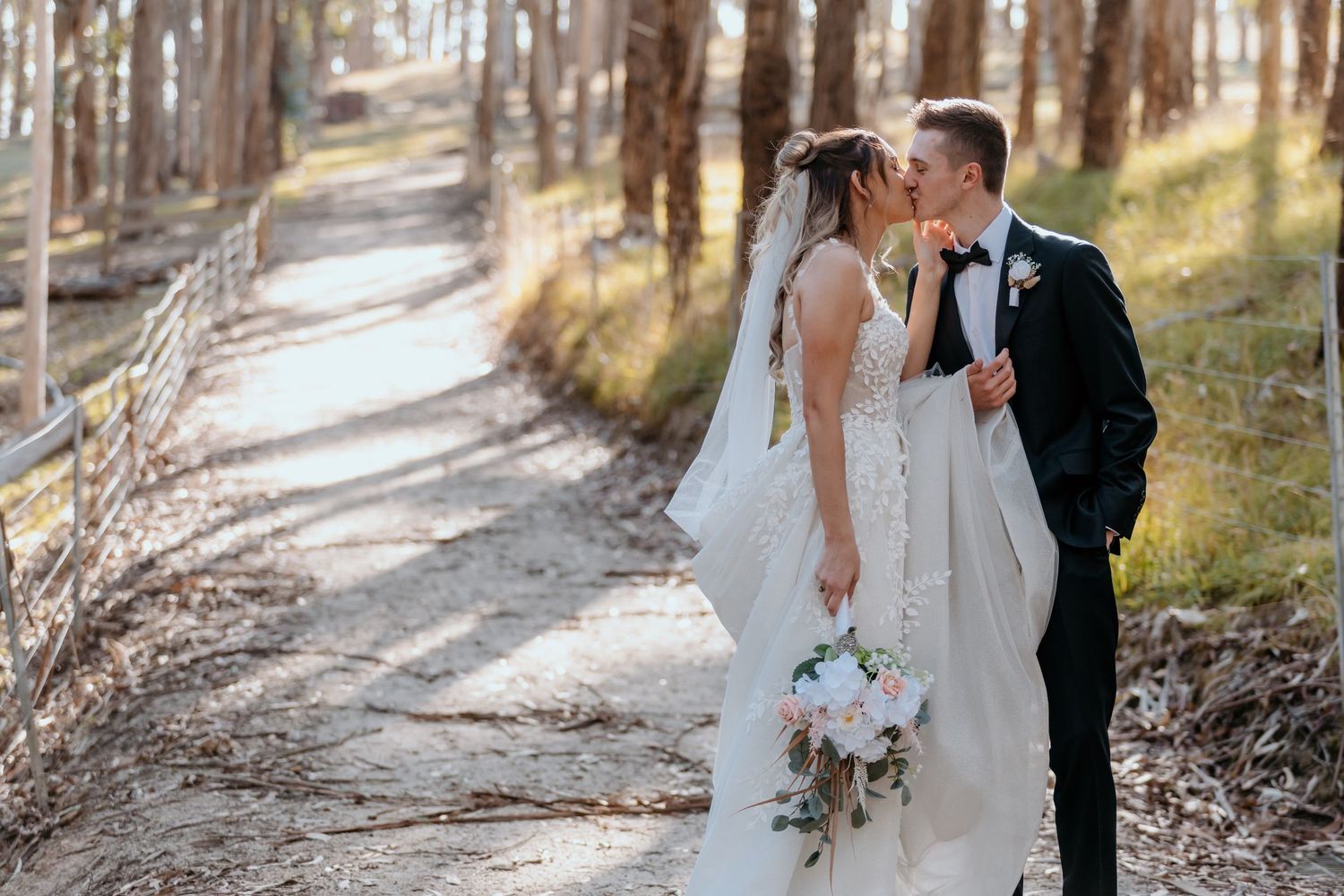 Wedding couple sharing intimate moment on tree-lined forest path with white bouquet.