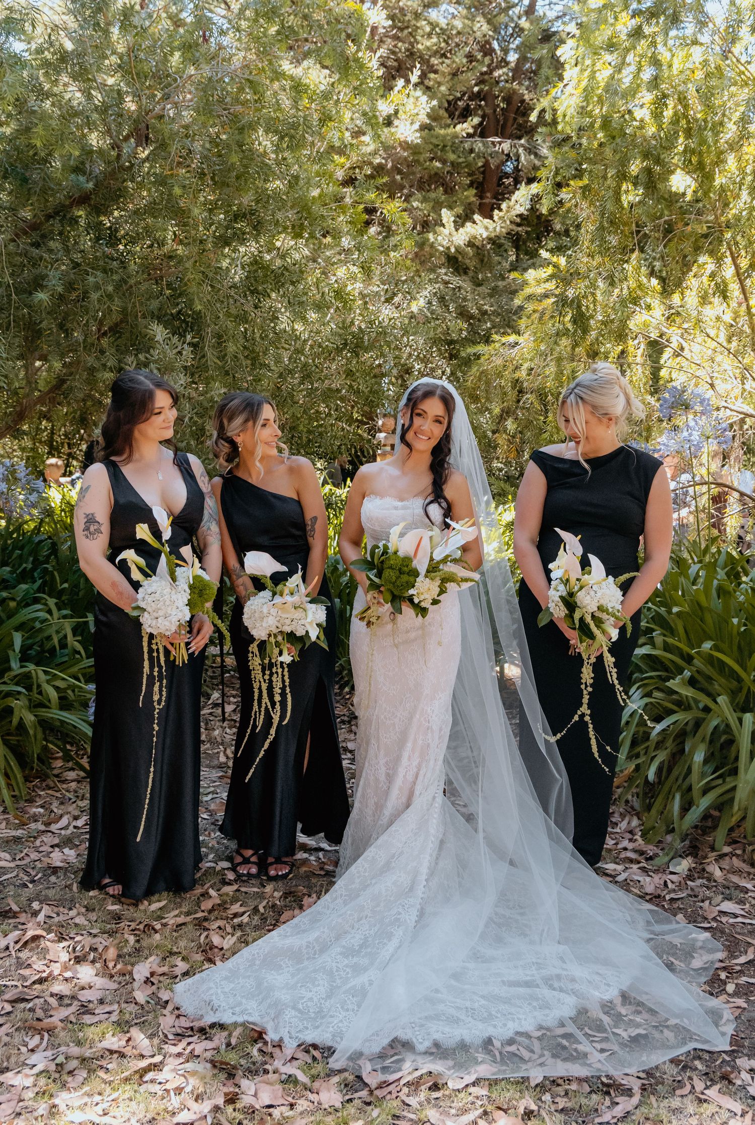 Bride in white lace gown with veil stands with three bridesmaids in black dresses holding white bouquets outdoors.