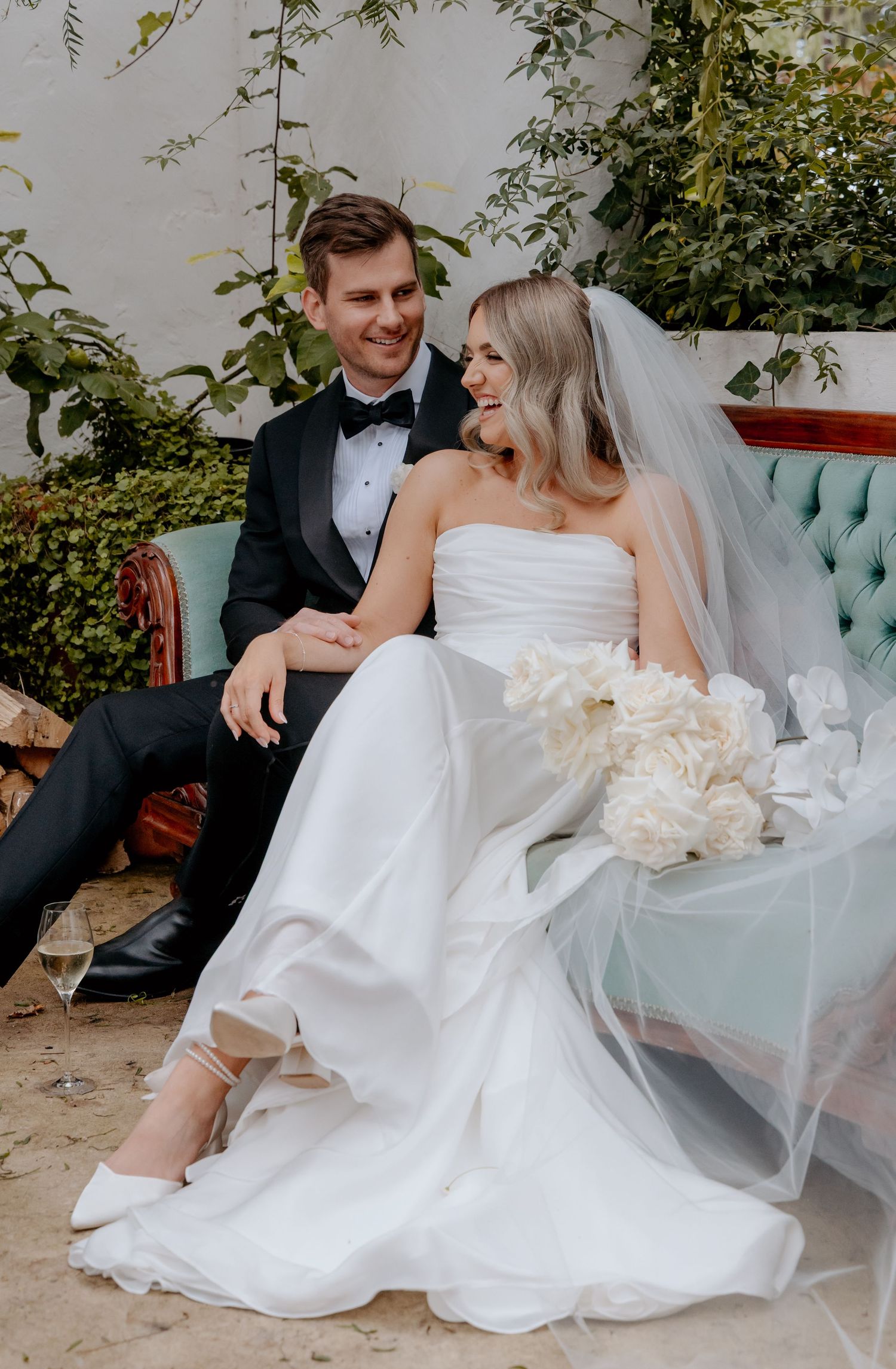 Bride and groom sitting together on a teal sofa, smiling, bride holding white bouquet in wedding gown and veil.