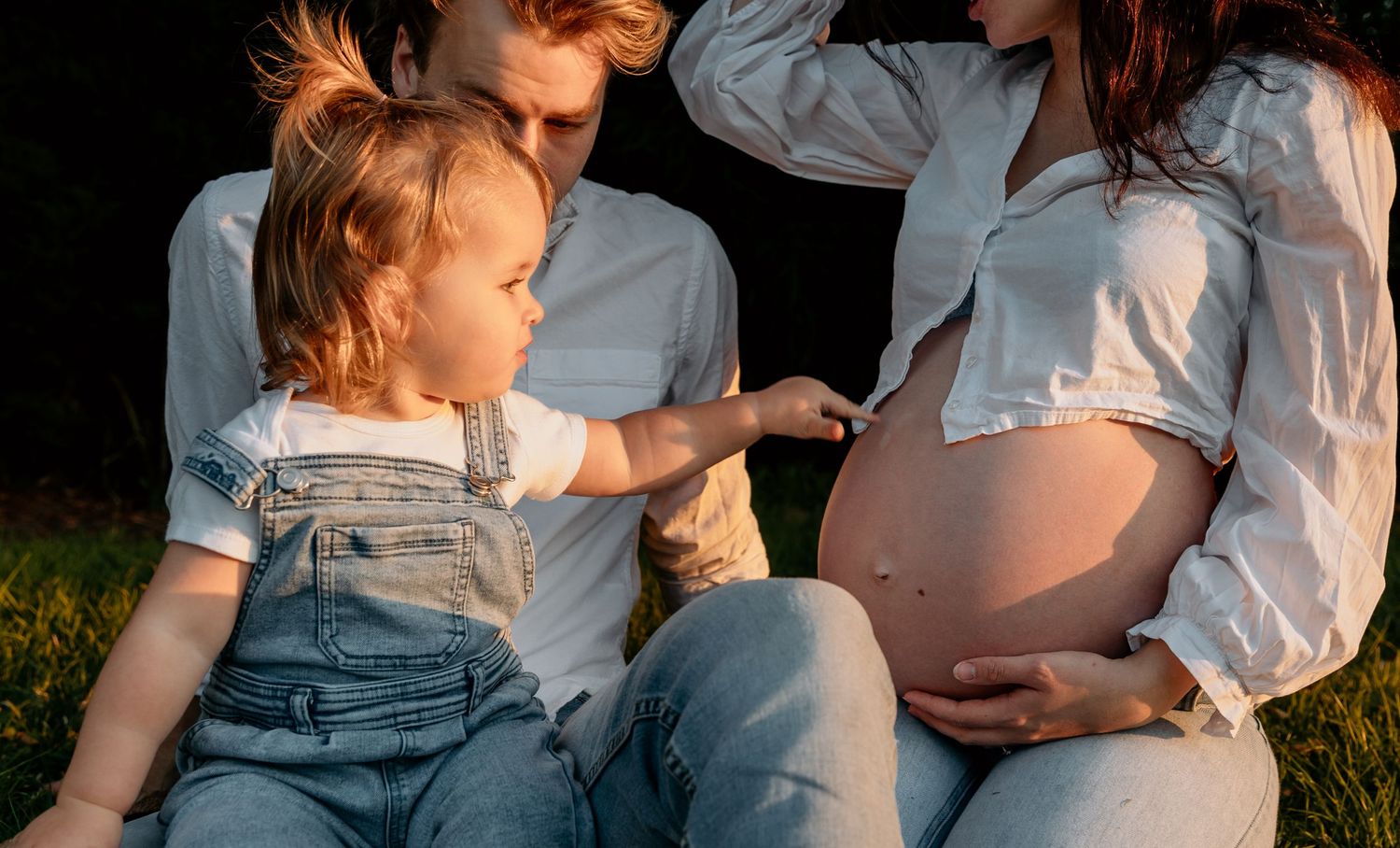 Child in overalls touches pregnant belly during outdoor family photo session.