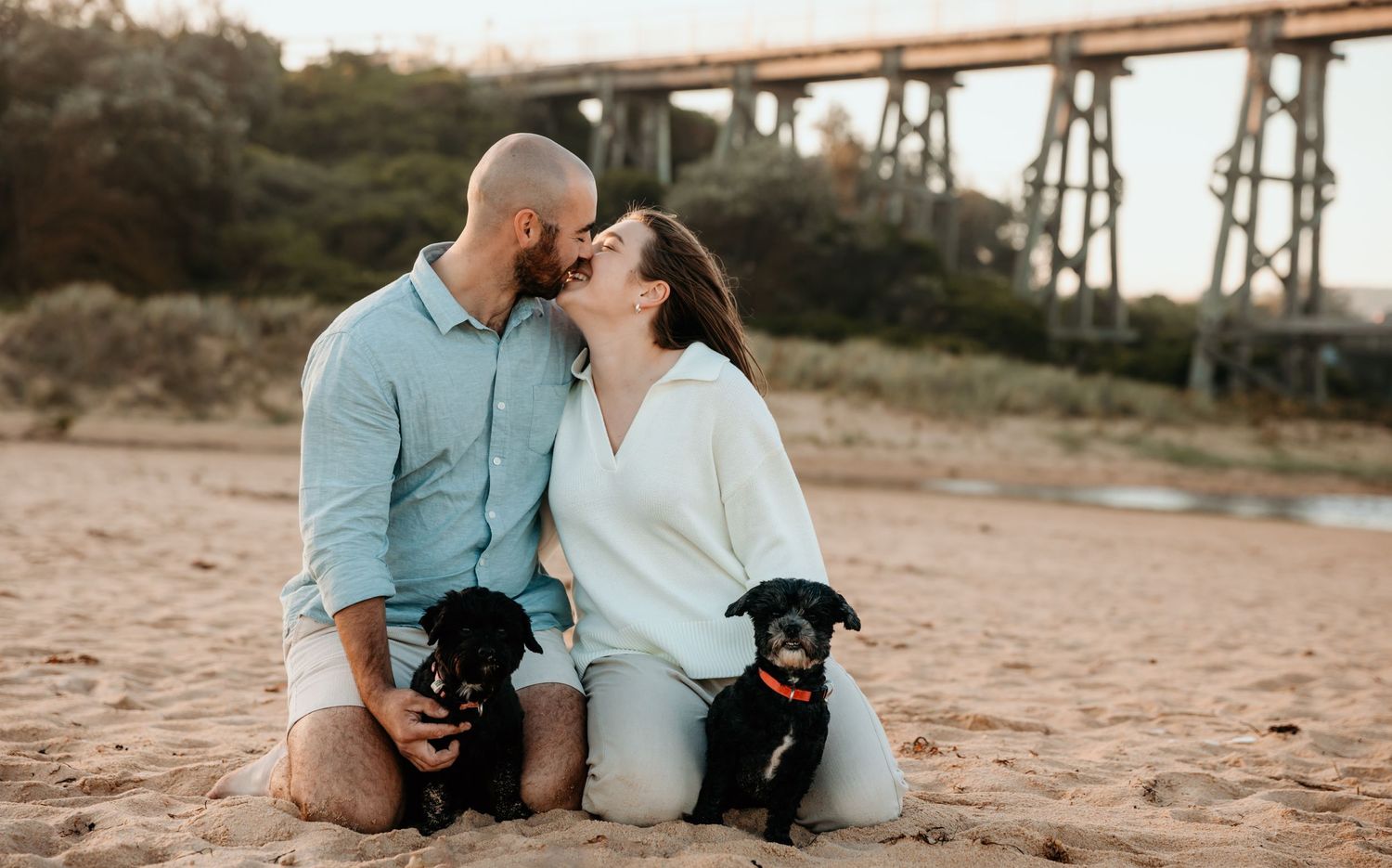Couple with two black dogs posing in front of rustic bridge at golden hour.
