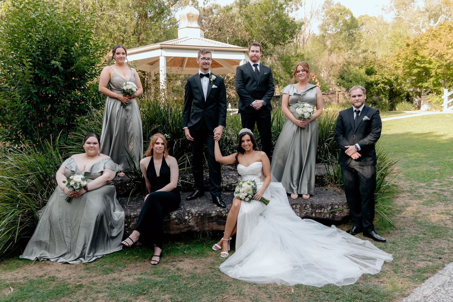 Wedding party members in gray bridesmaid dresses and dark suits pose together on grassy lawn at outdoor ceremony.