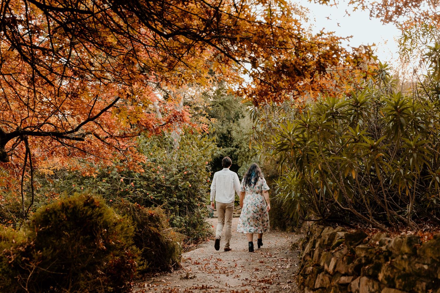 A couple walks hand in hand down a leaf-covered path beneath autumn trees.