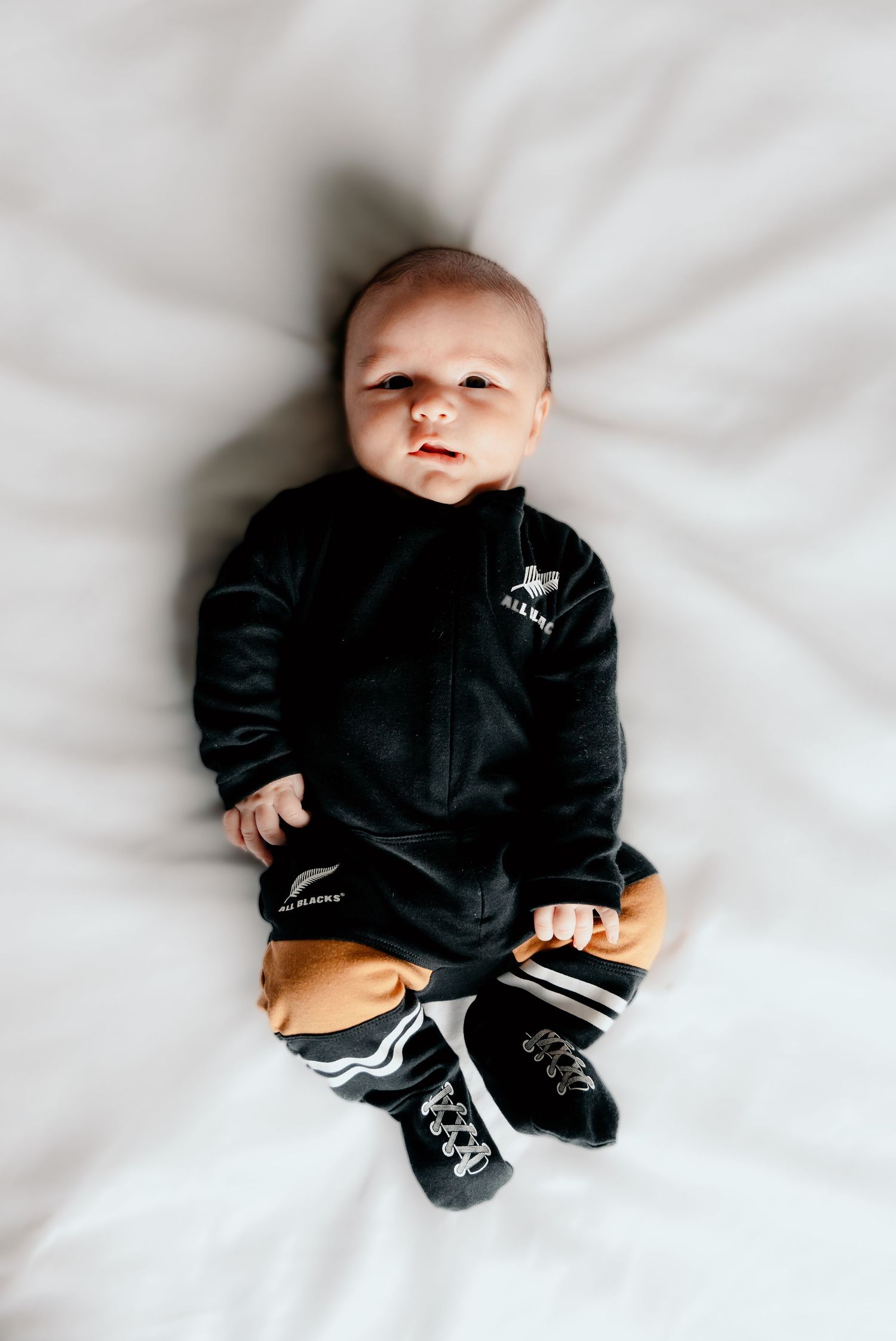 A baby wearing a black onesie lies on white bedding in three different poses.