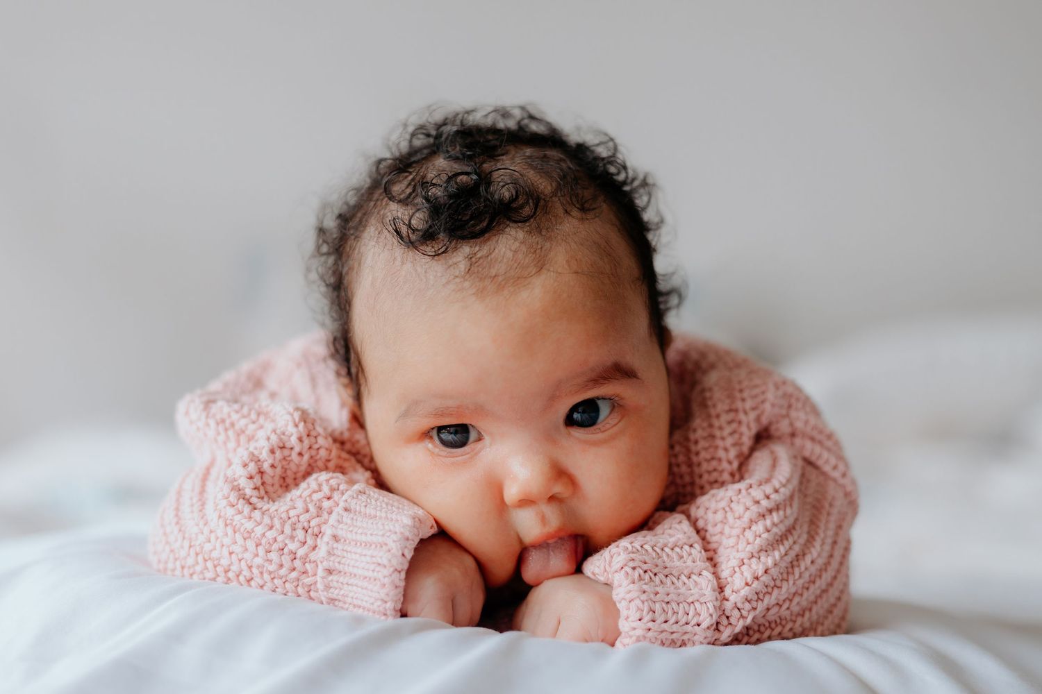 Newborn baby wearing soft pink knit clothing resting peacefully on white bedding.