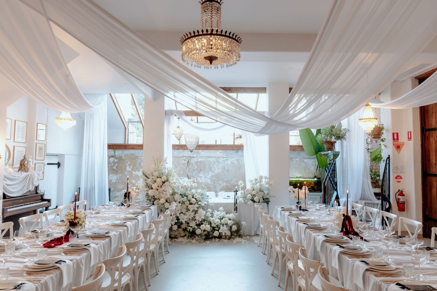 Elegant white wedding reception hall with draped ceilings, chandelier, floral arrangements, and neatly set dining tables.