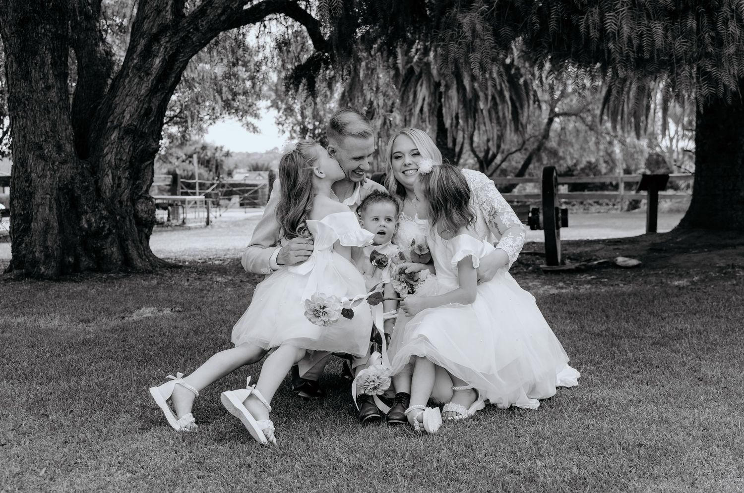 Black and white photo of a couple in wedding attire hugging two young girls in white dresses under a large tree.