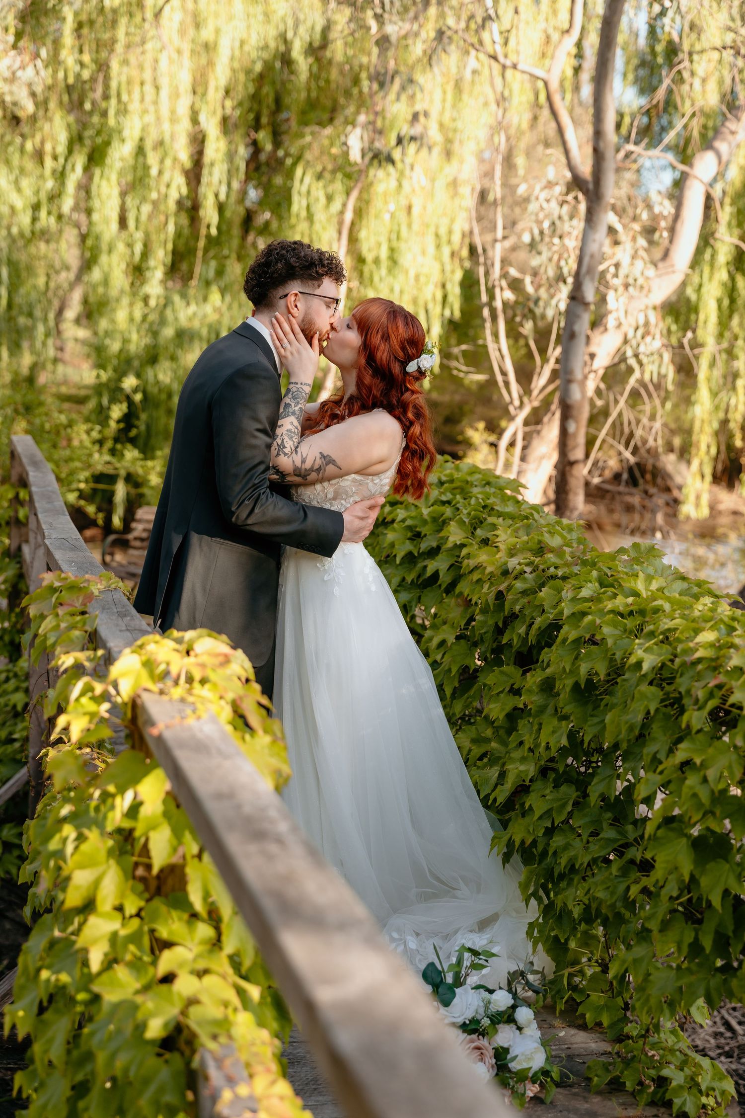 A romantic wedding couple shares a tender embrace in a garden setting with sunlit willow trees and lush greenery.