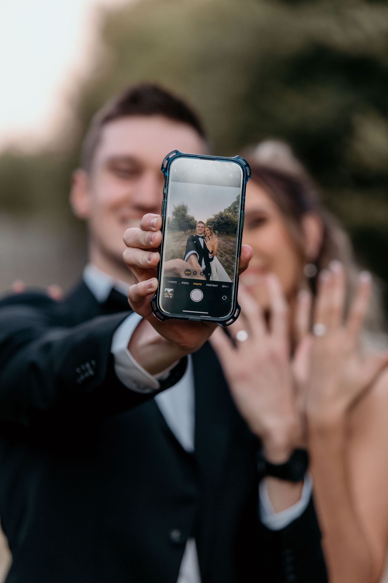 A hand holds up a phone displaying a photo while a couple shows off an engagement ring in the background.