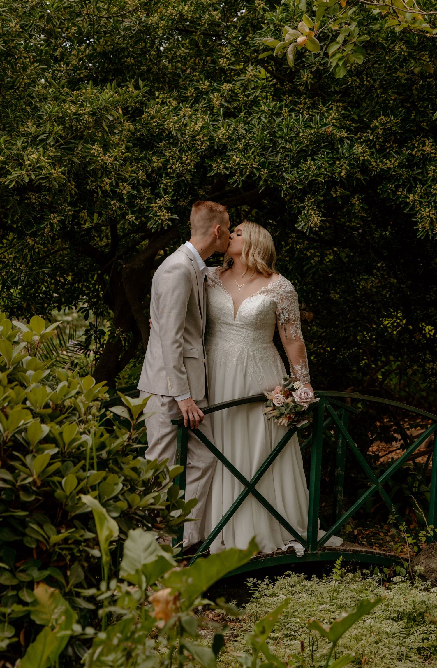 Bride and groom share a romantic kiss on a green garden bridge framed by dense foliage.