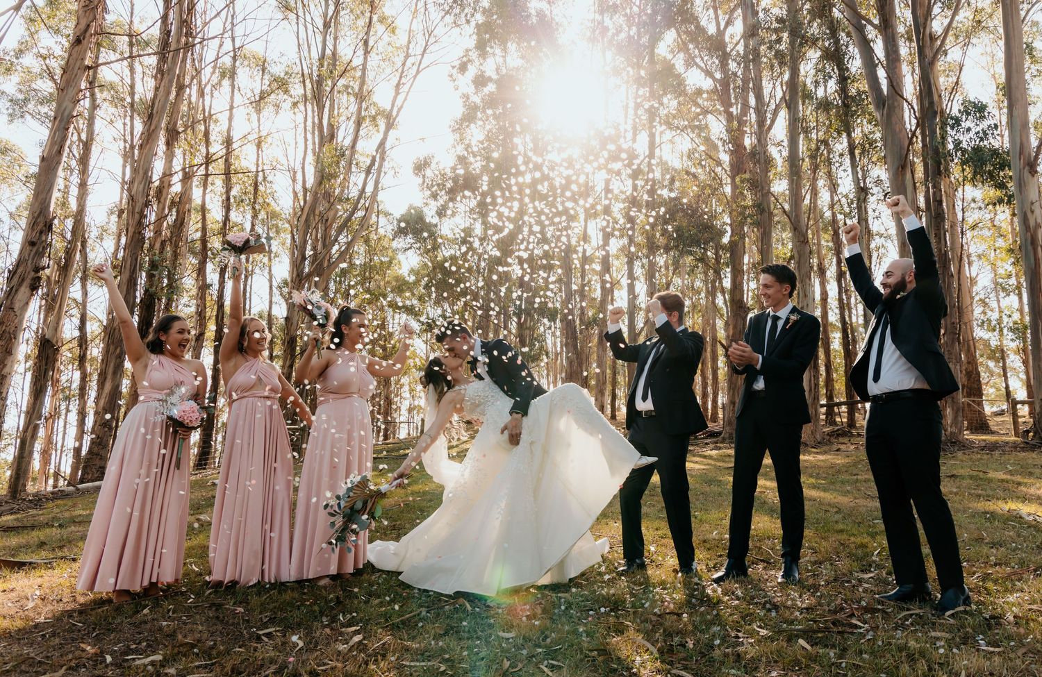 Wedding party celebrates in sunlit forest with bridesmaids in pink dresses and groomsmen in suits dancing together.