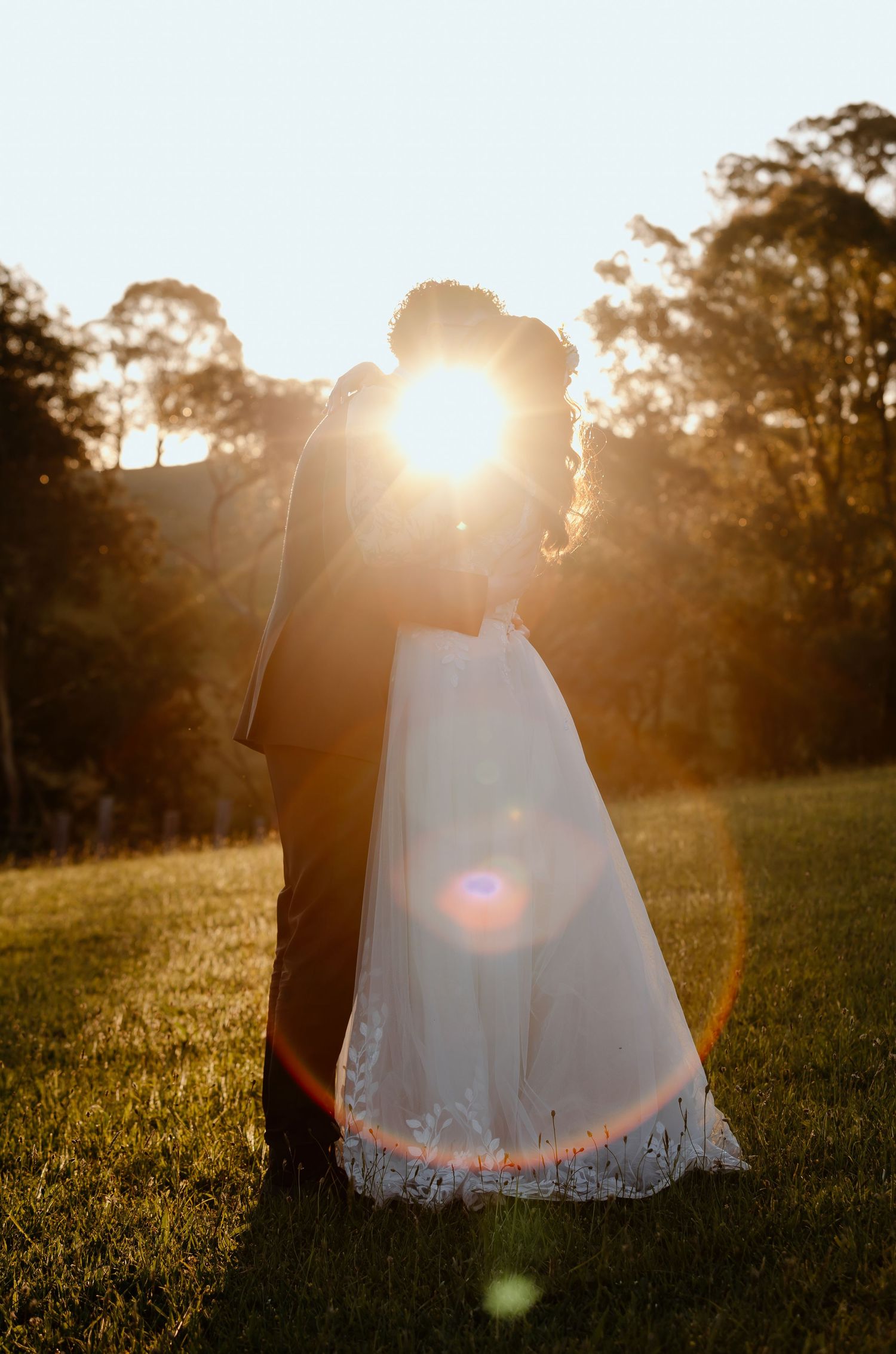 Romantic sunset silhouettes of newlyweds in wedding attire standing on grassy hill with lens flare effect.