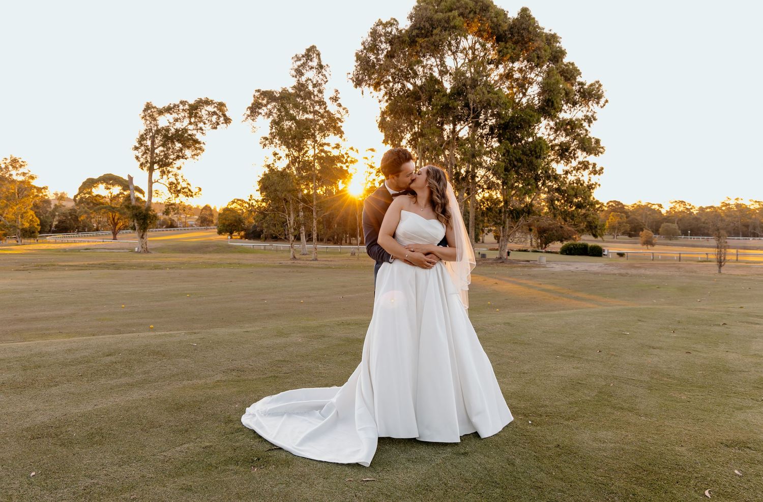 A bride in an elegant white gown stands in a grassy field at sunset with trees in the background.