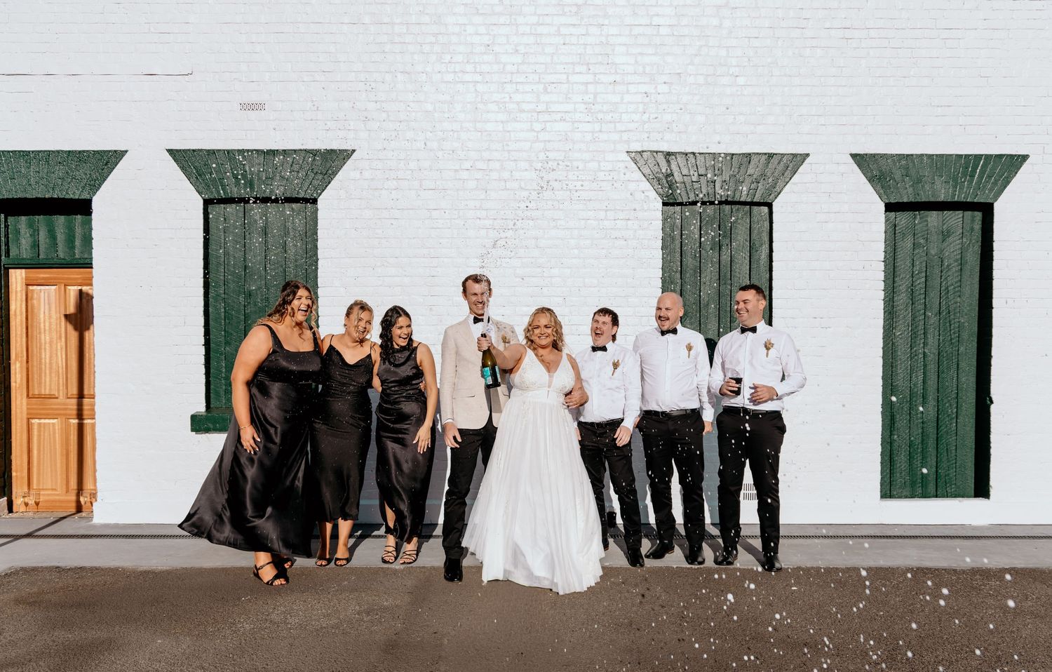 Wedding party poses along white brick wall with green shutters and wooden doors at outdoor venue.