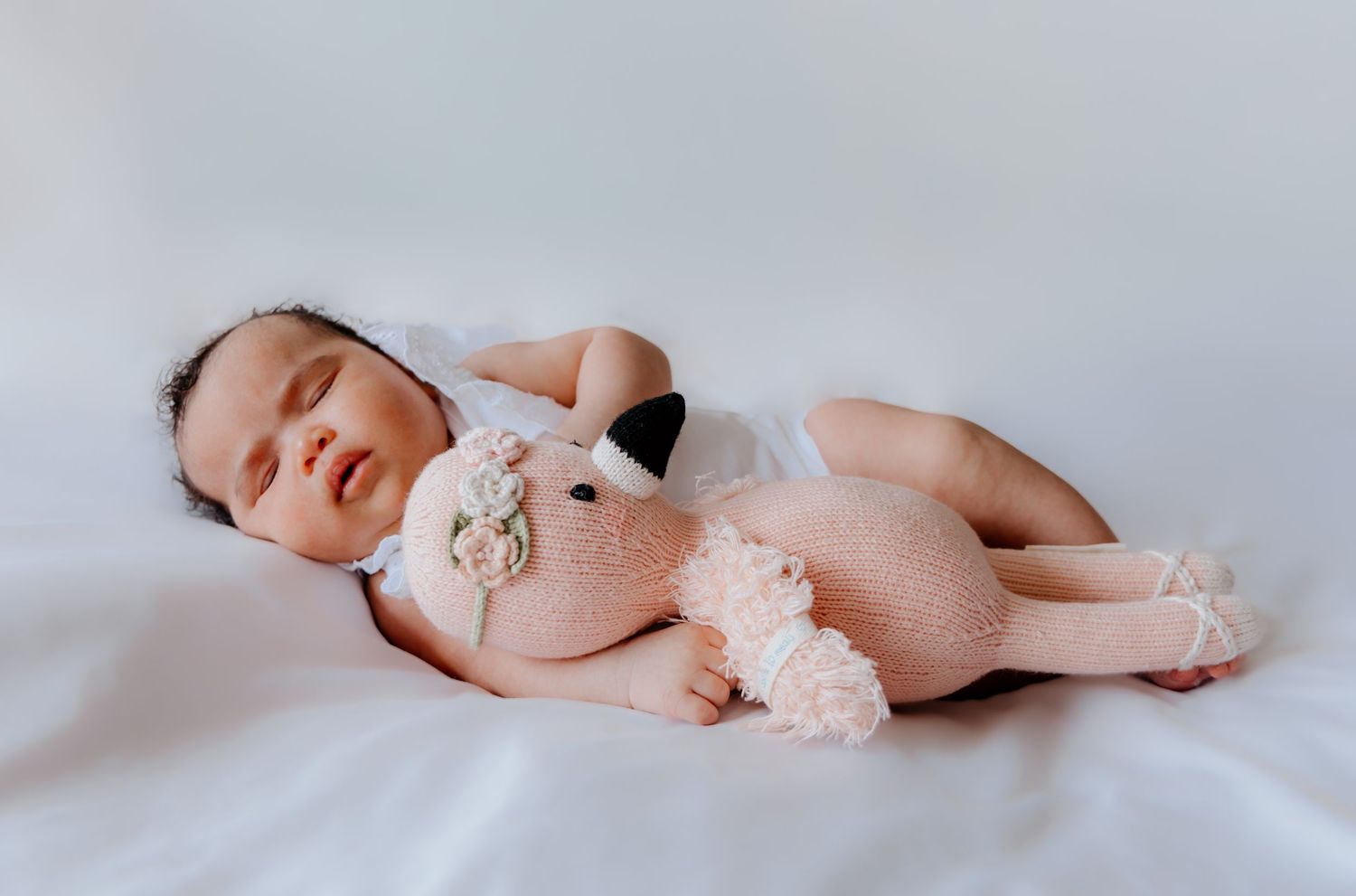 Sleeping baby wearing a pink onesie with angel wings lies peacefully on white bedding.