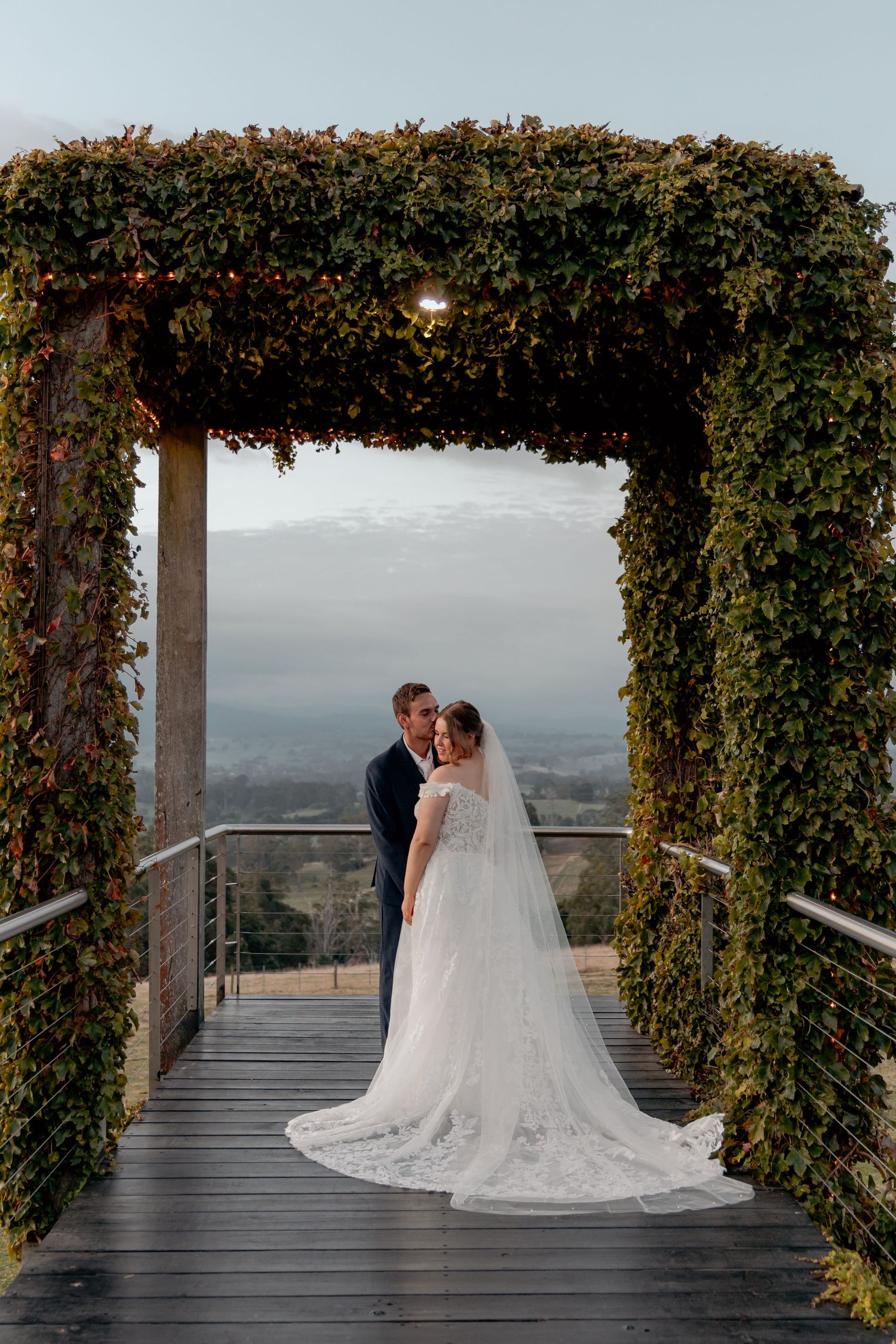 A romantic wedding couple embraces on a scenic mountain overlook deck surrounded by lush green vines and foliage.
