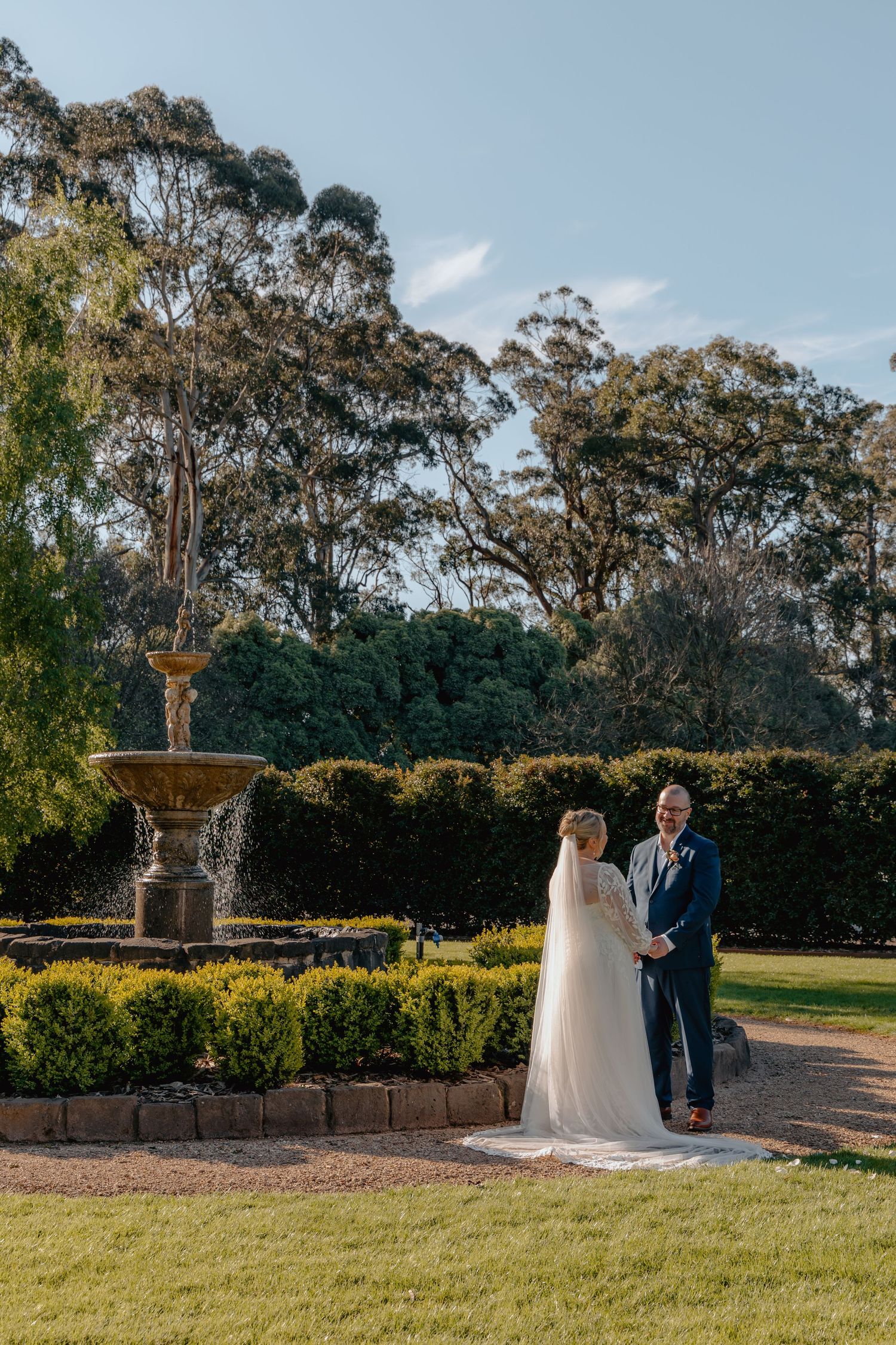 A romantic wedding couple stands near a decorative fountain in a garden setting with tall eucalyptus trees in the background.