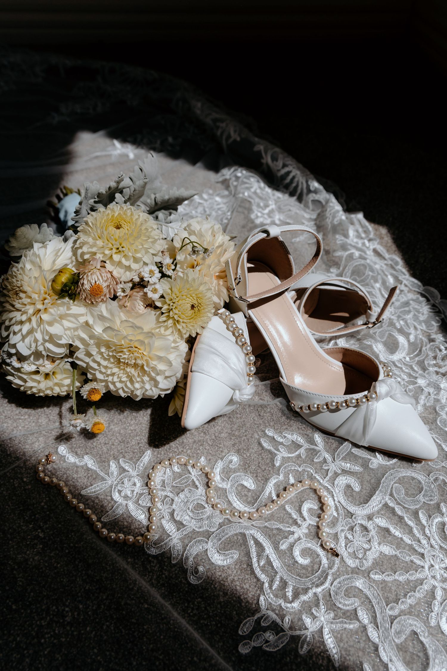 White bridal heels with pearl details beside a bouquet of cream dahlias and a pearl necklace on lace fabric.