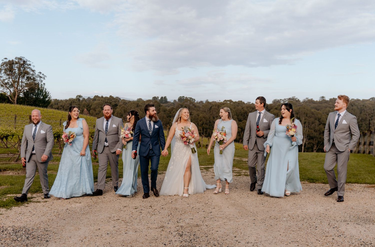 Wedding party in light blue and gray attire poses together on a rustic outdoor path.