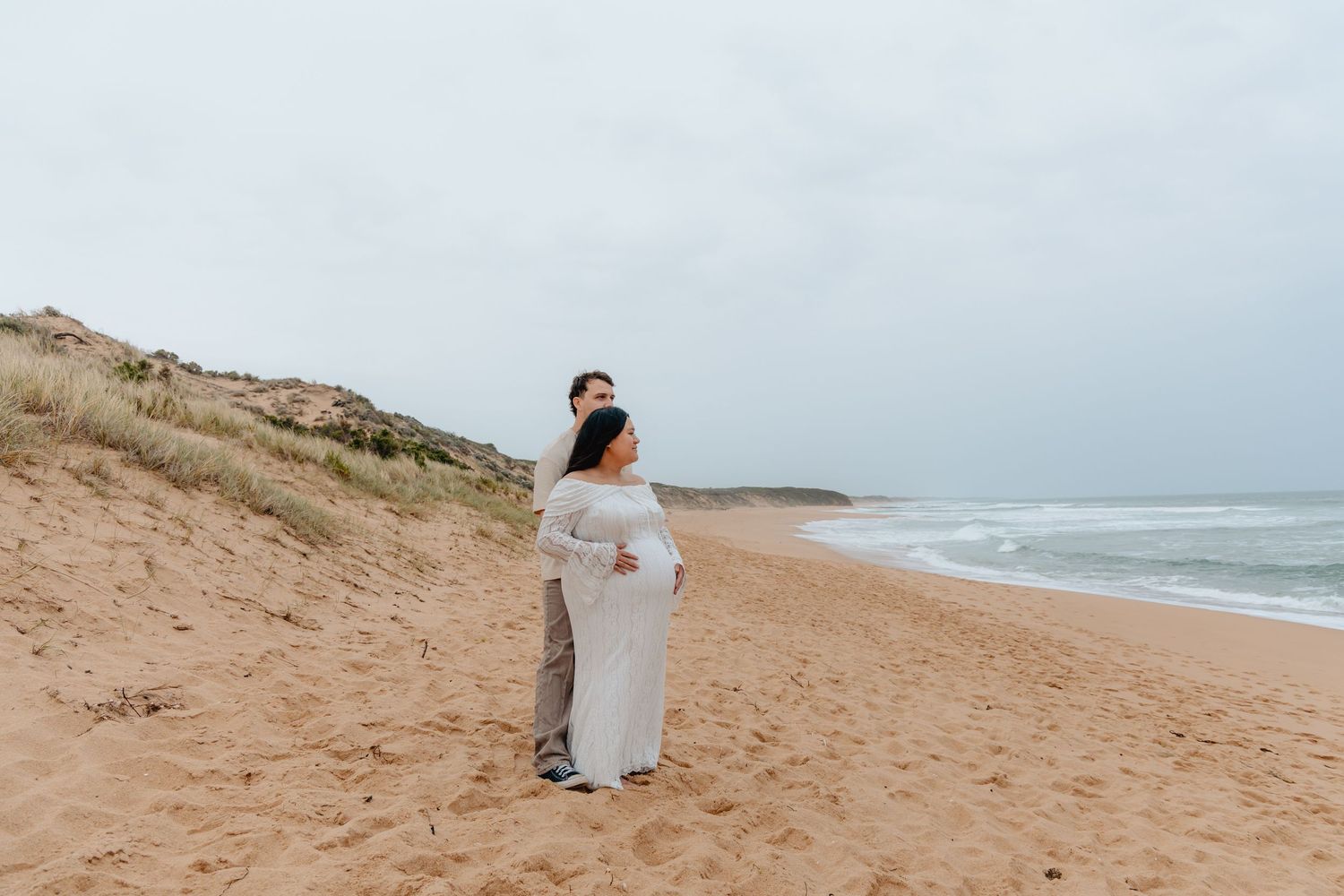 A couple in white attire walks along a sandy beach with ocean waves crashing in the background.