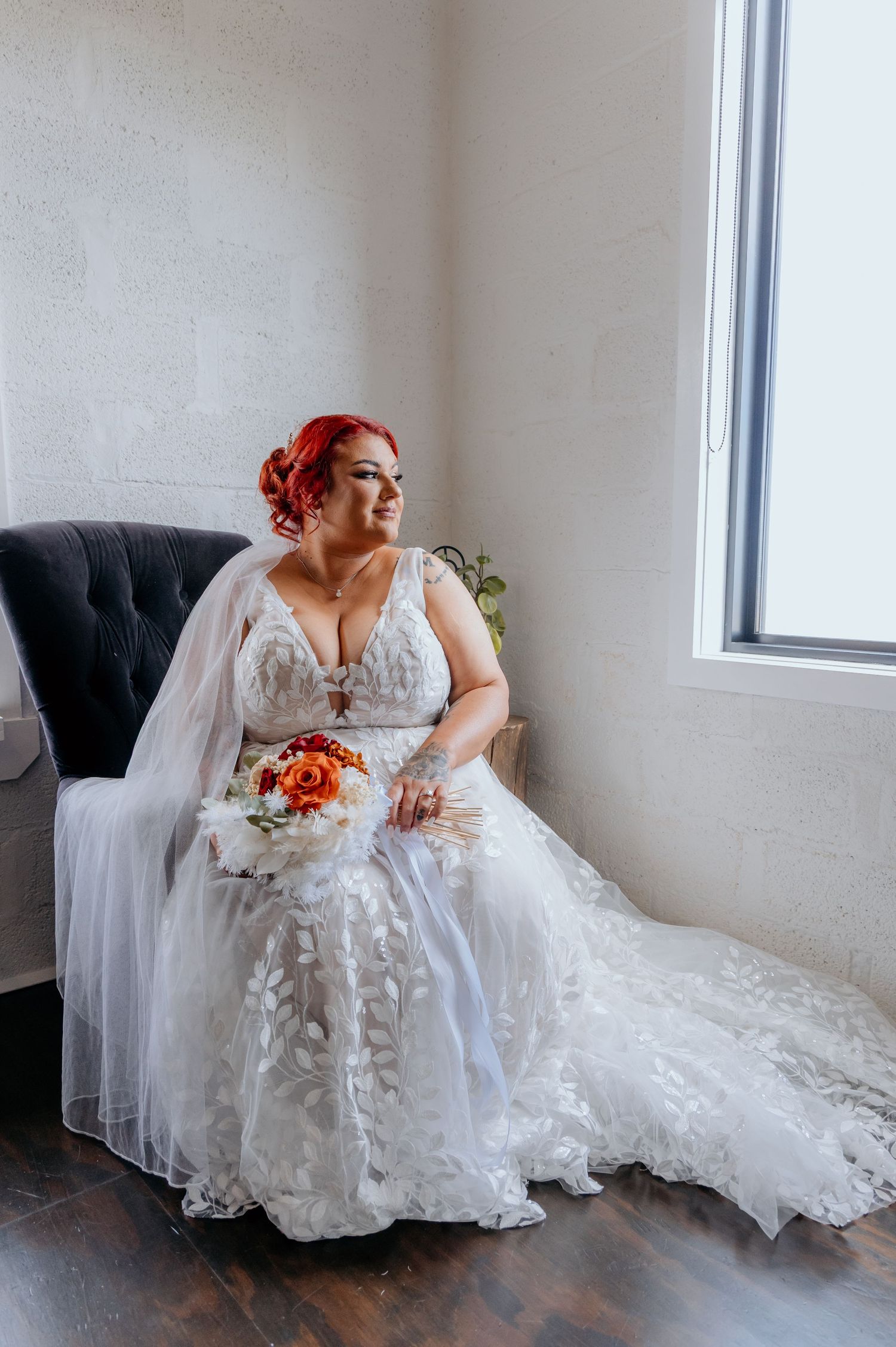 A bride with red hair in a white lace wedding gown sits in a chair holding an orange bouquet near a bright window.