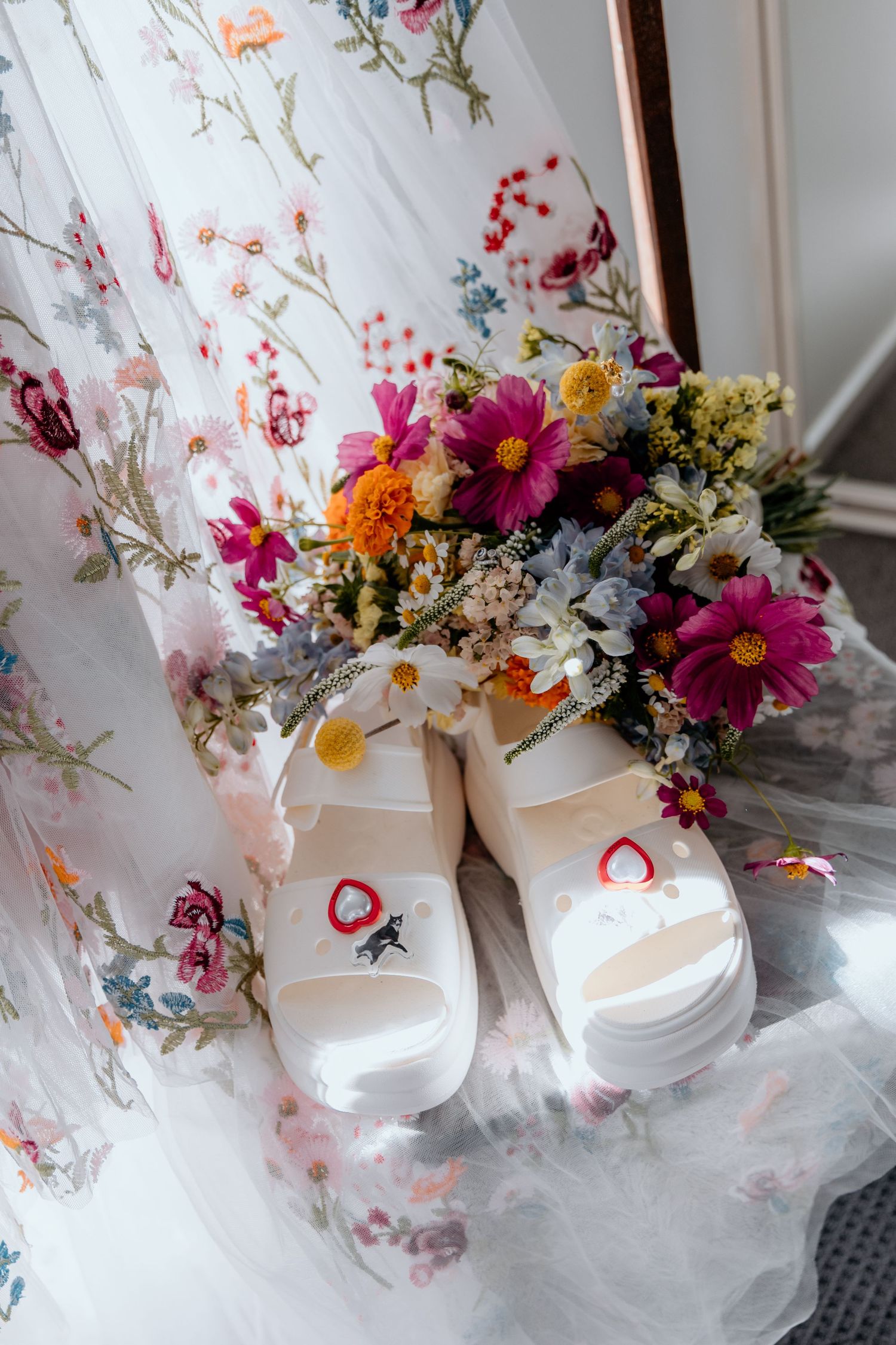 White Crocs with heart Jibbitz sit beside a colorful wildflower bouquet on a floral wedding dress.