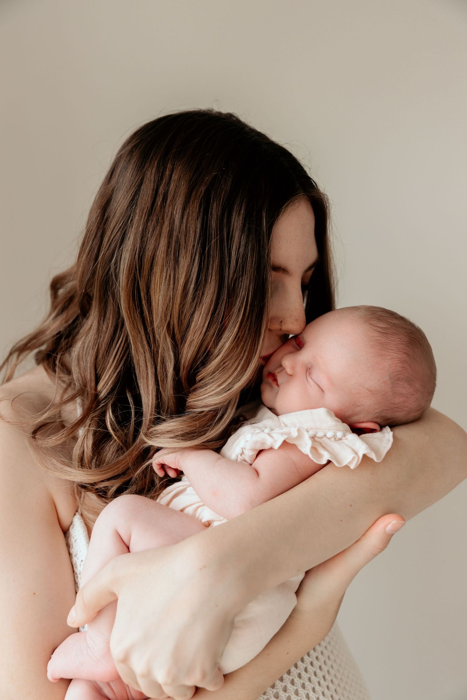 A mother with ombre hair tenderly kisses her newborn baby against a neutral background.