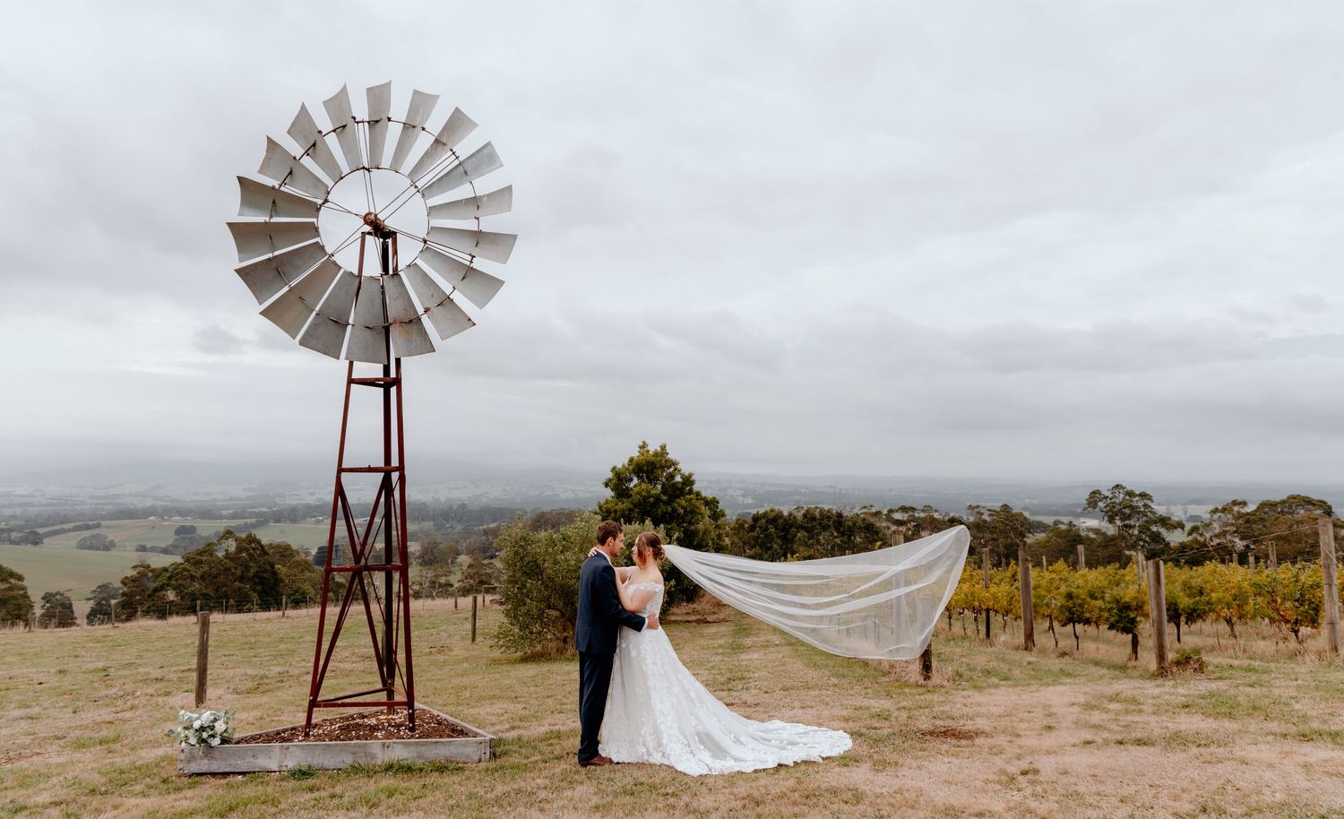 A rural countryside wedding ceremony in front of a rustic windmill with a flowing veil caught in the breeze.