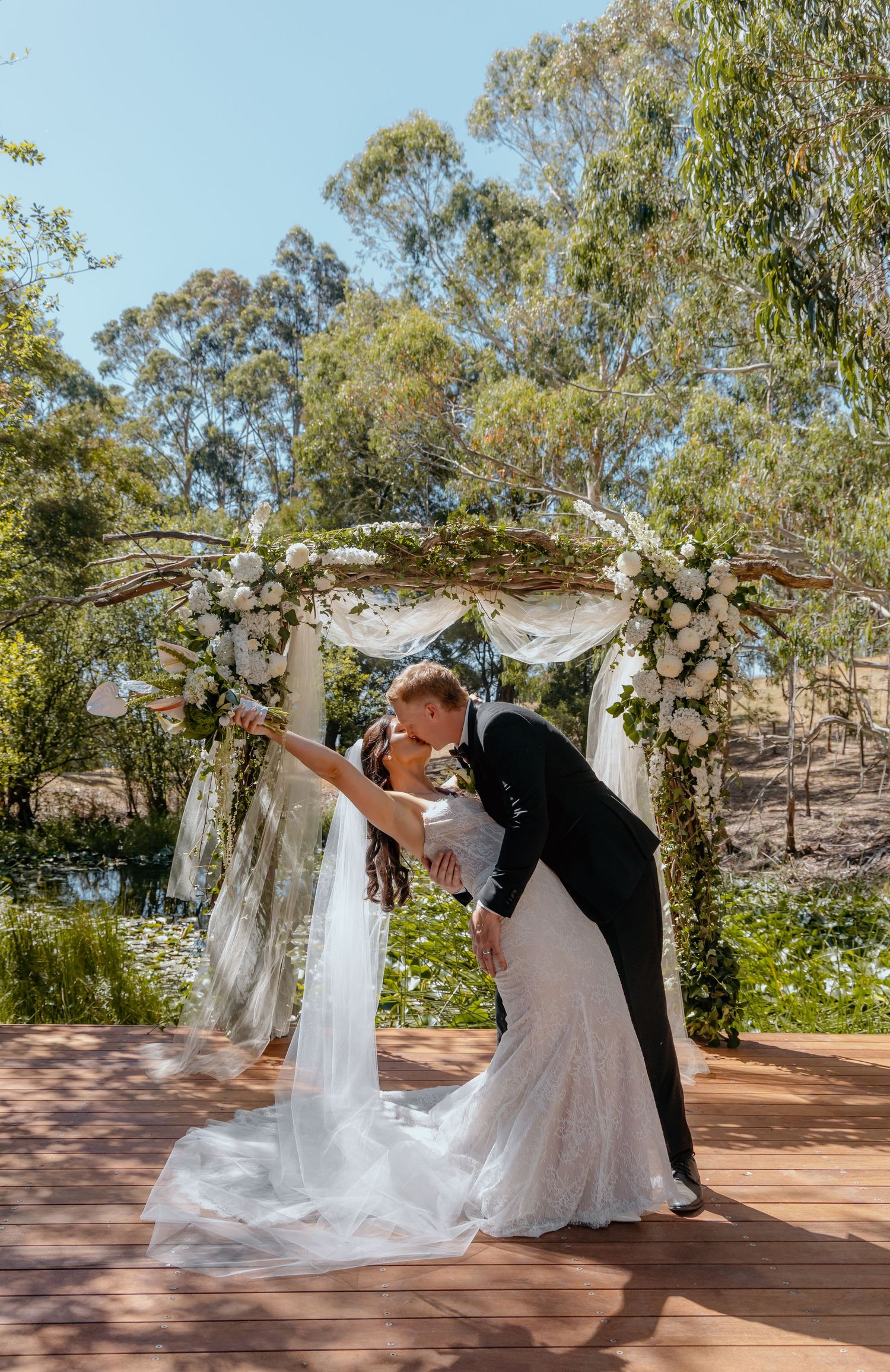 Bride and groom share a romantic dip kiss under a floral arch adorned with white roses and draped fabric outdoors.