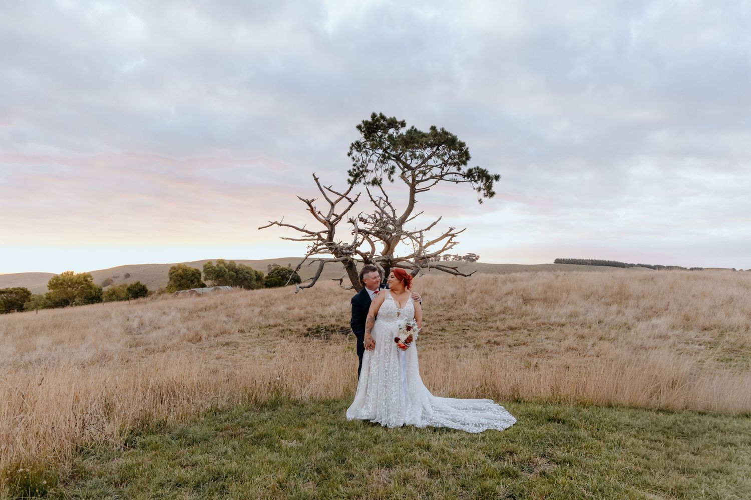 Bride and groom standing in golden field beside bare tree at sunset during outdoor wedding ceremony.